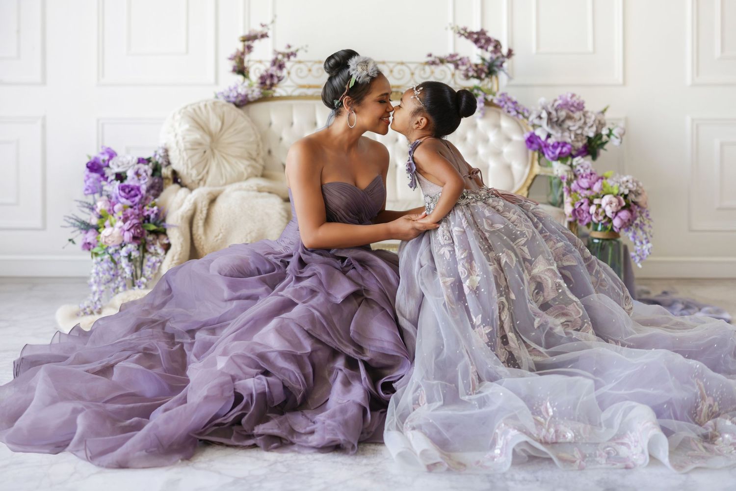 Mother and daughter in matching purple tulle gowns share a tender kiss during an elegant studio portrait session.
