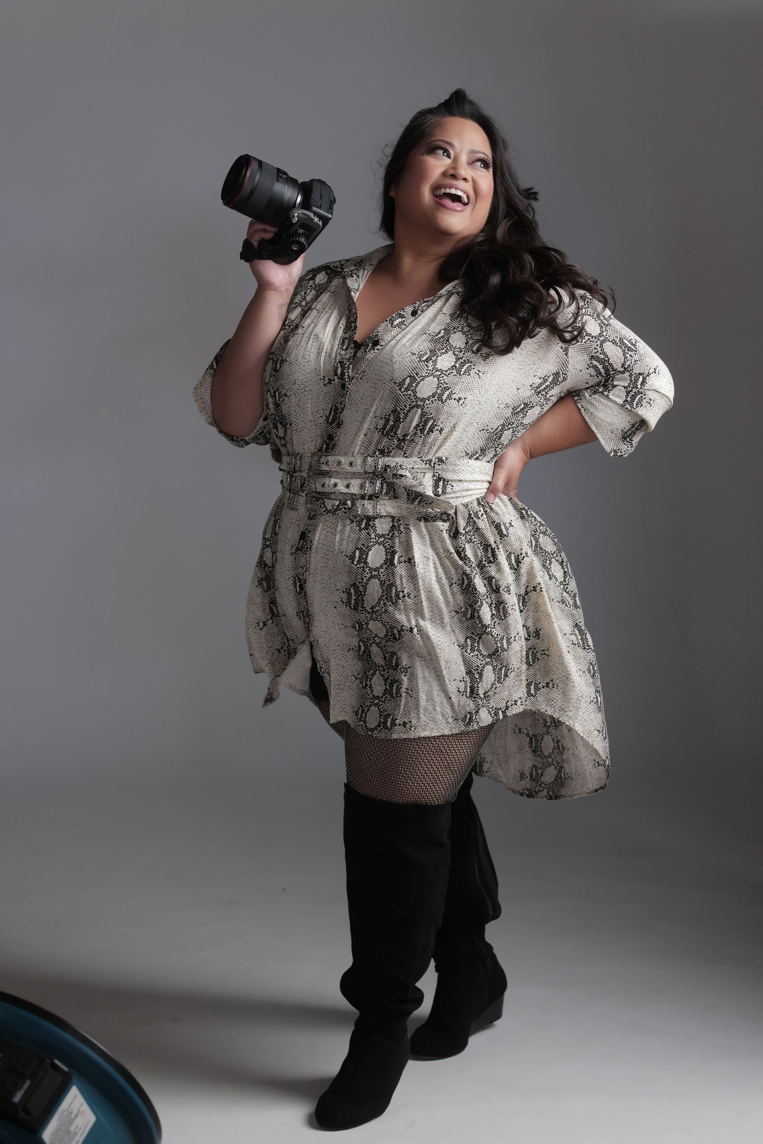 Plus-size woman in snakeskin print dress and knee-high boots poses confidently holding a professional camera in studio.
