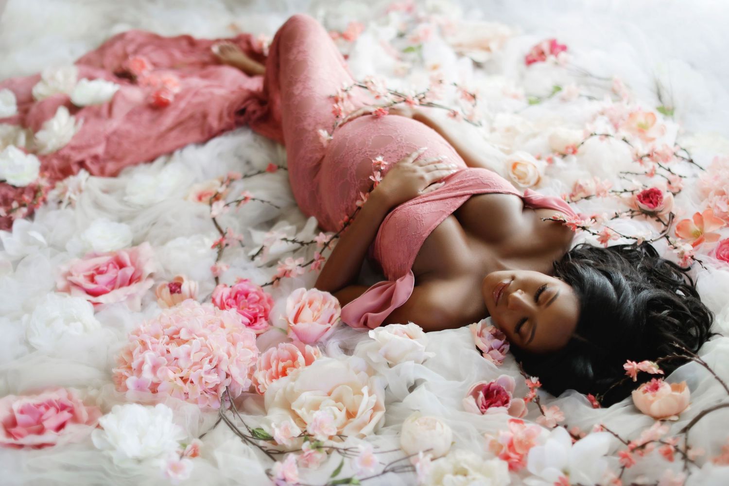 Pregnant woman in pink maternity outfit lying among pink and white roses for a floral maternity photoshoot.
