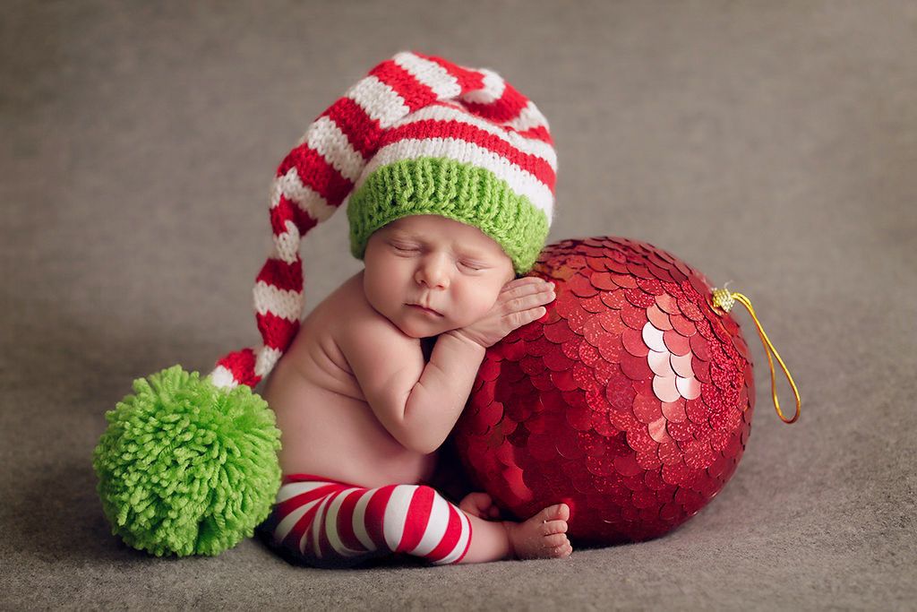 Sleeping newborn baby wearing a red and white striped Christmas elf hat and leggings, cuddled next to a large red ornament.