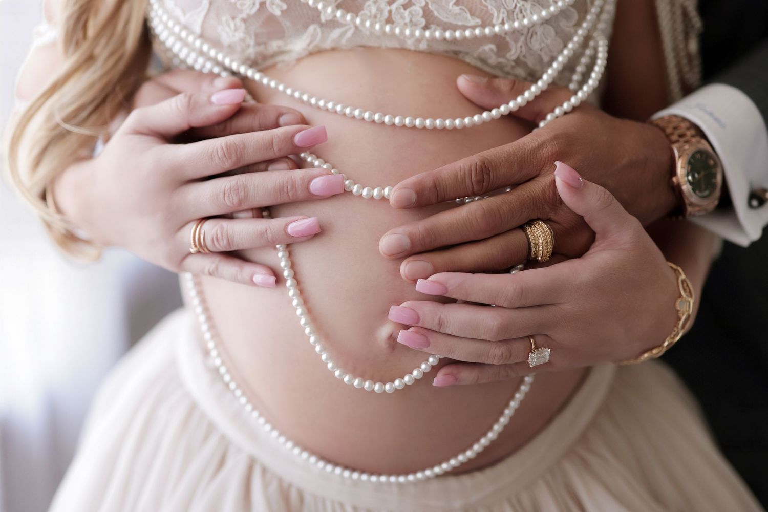 Pregnant belly adorned with draped pearl necklaces, hands gently holding bump.