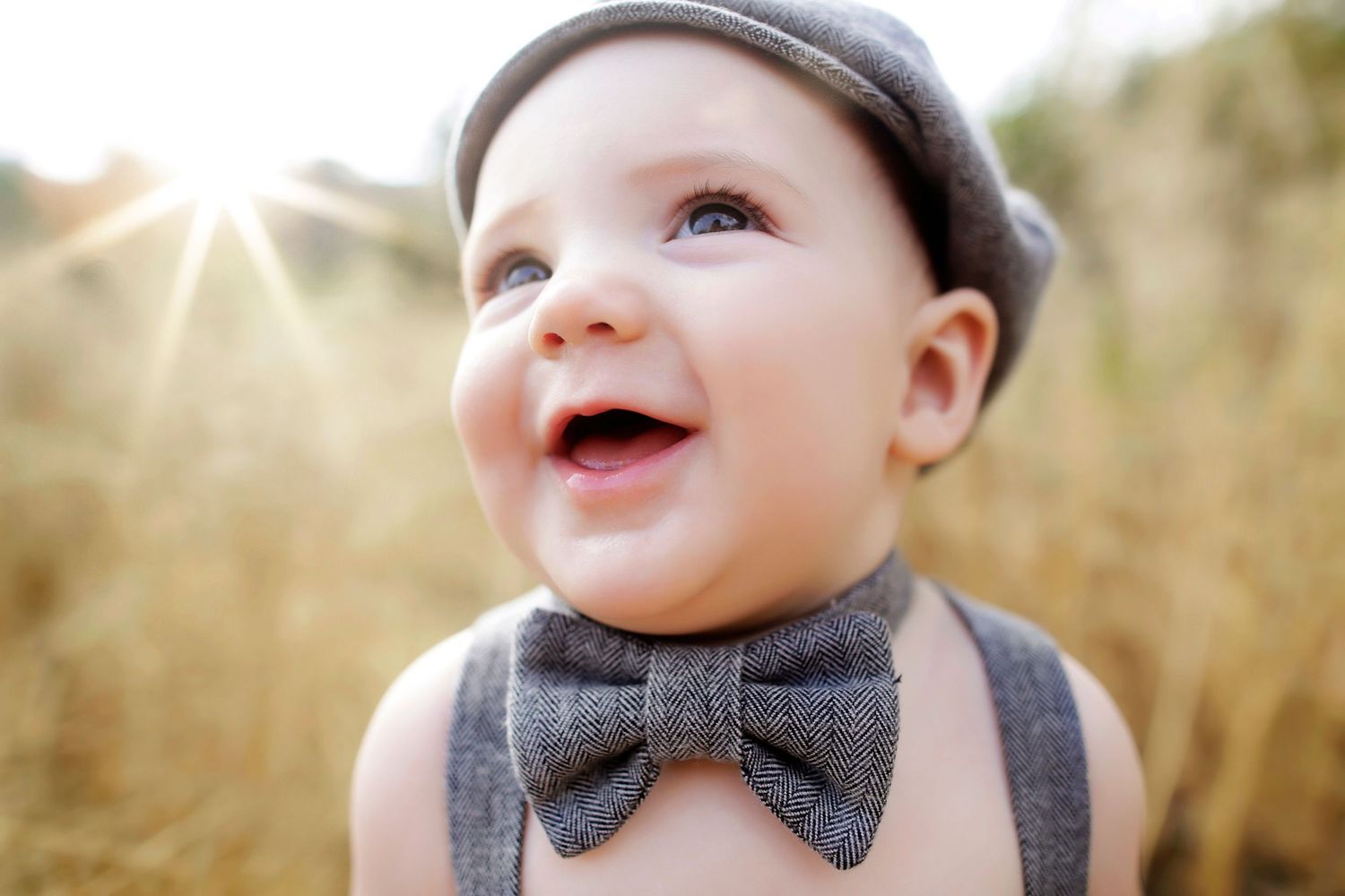 Adorable smiling baby wearing a gray flat cap and large bow tie with suspenders, bathed in warm sunlight outdoors.