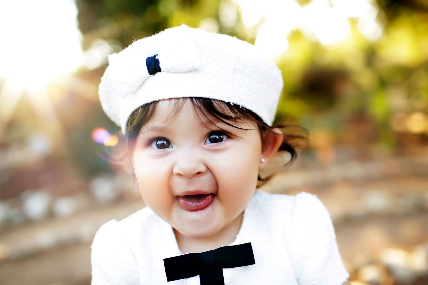 Adorable smiling baby wearing a white hat and bow tie outfit, with bright eyes and chubby cheeks outdoors.