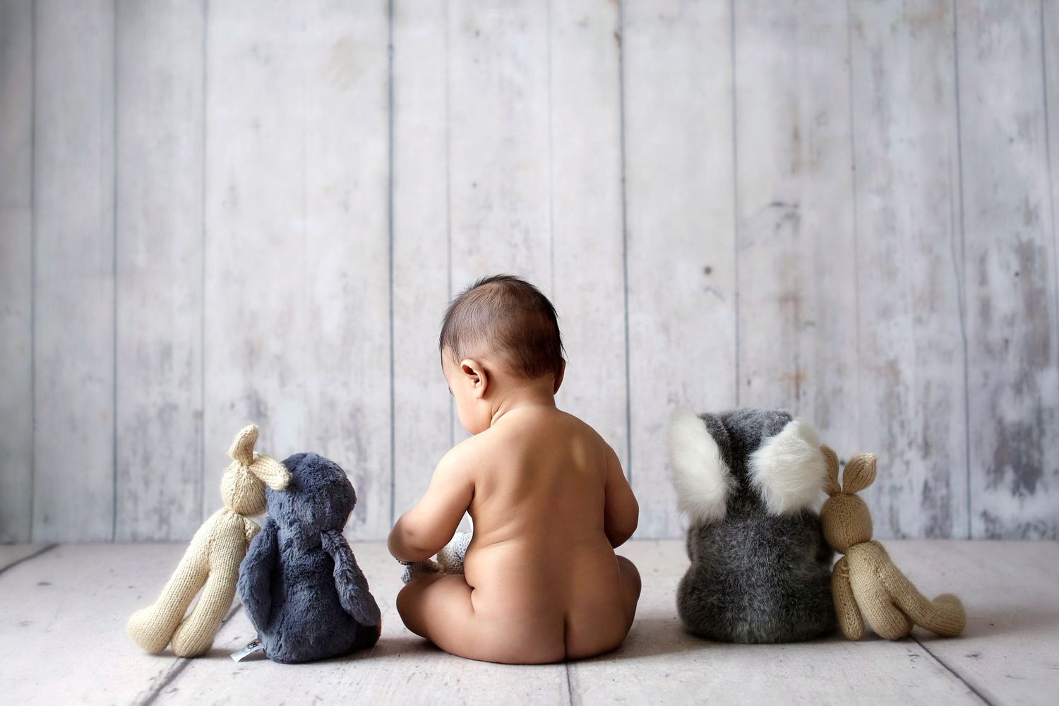 Baby sitting on floor facing away with four stuffed animal toys against a rustic wall.