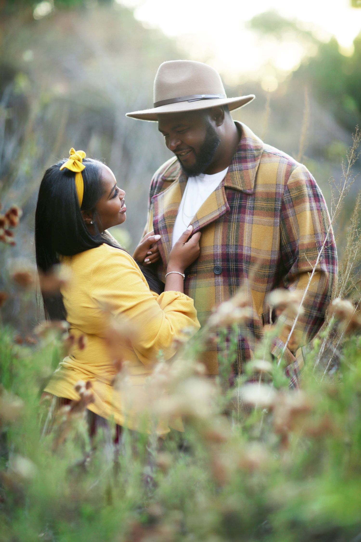 A couple shares a romantic moment in a sunlit field, woman in yellow dress touching man's plaid shirt.