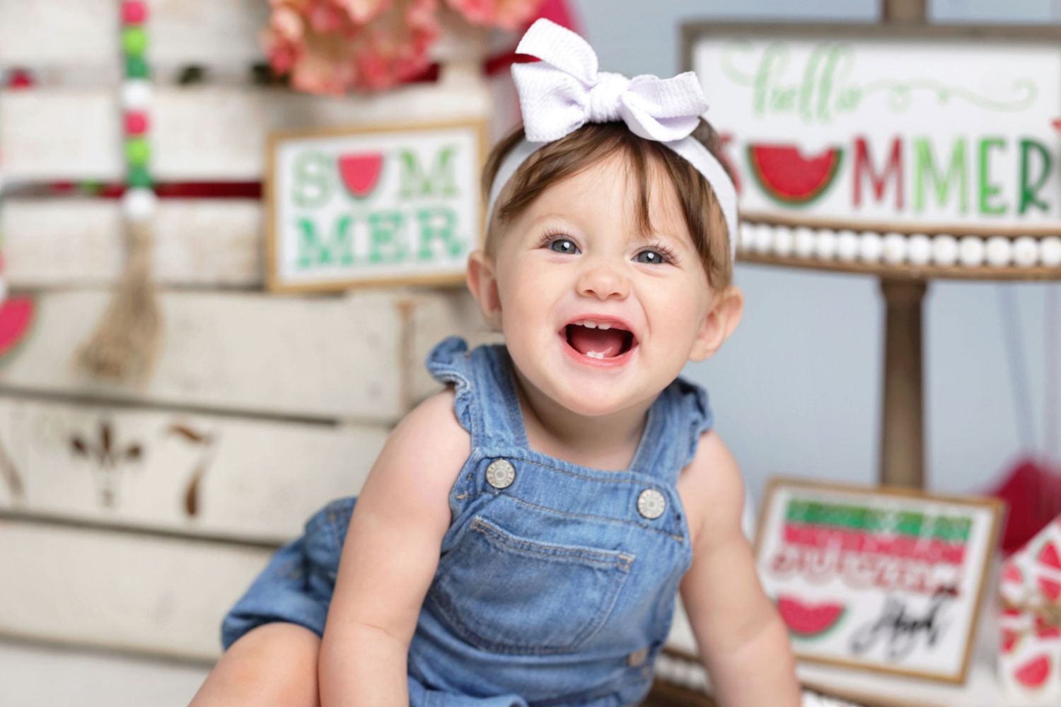 Laughing toddler girl in denim overalls with white bow headband in front of rustic summer and watermelon-themed signs.