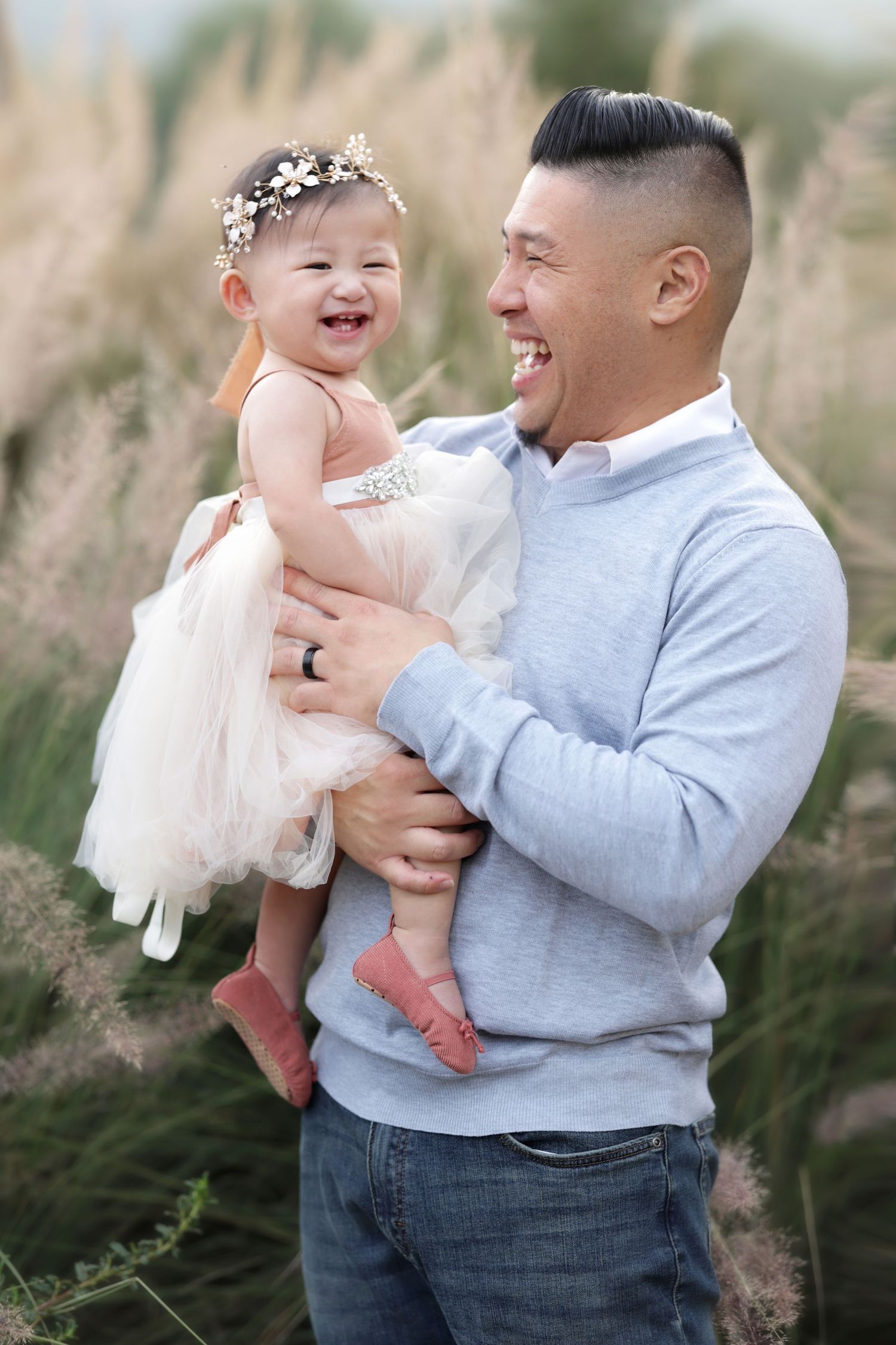 A smiling man holds a laughing baby girl in a white tutu dress outdoors in a field of tall grasses.