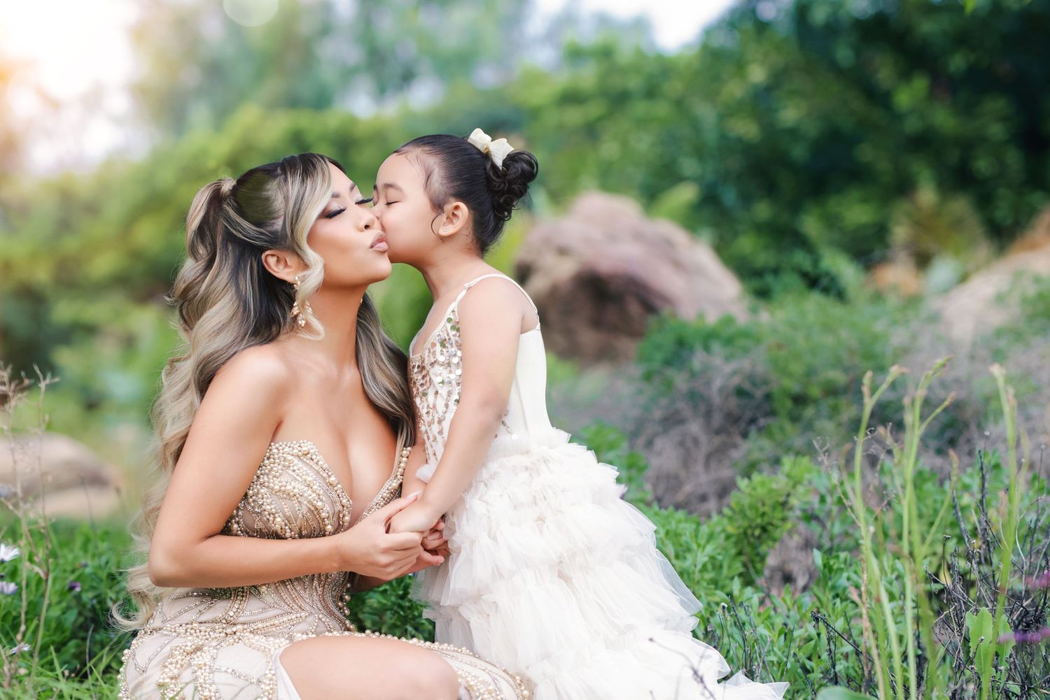 A woman in a gold gown shares a kiss with a young girl in a white dress outdoors.