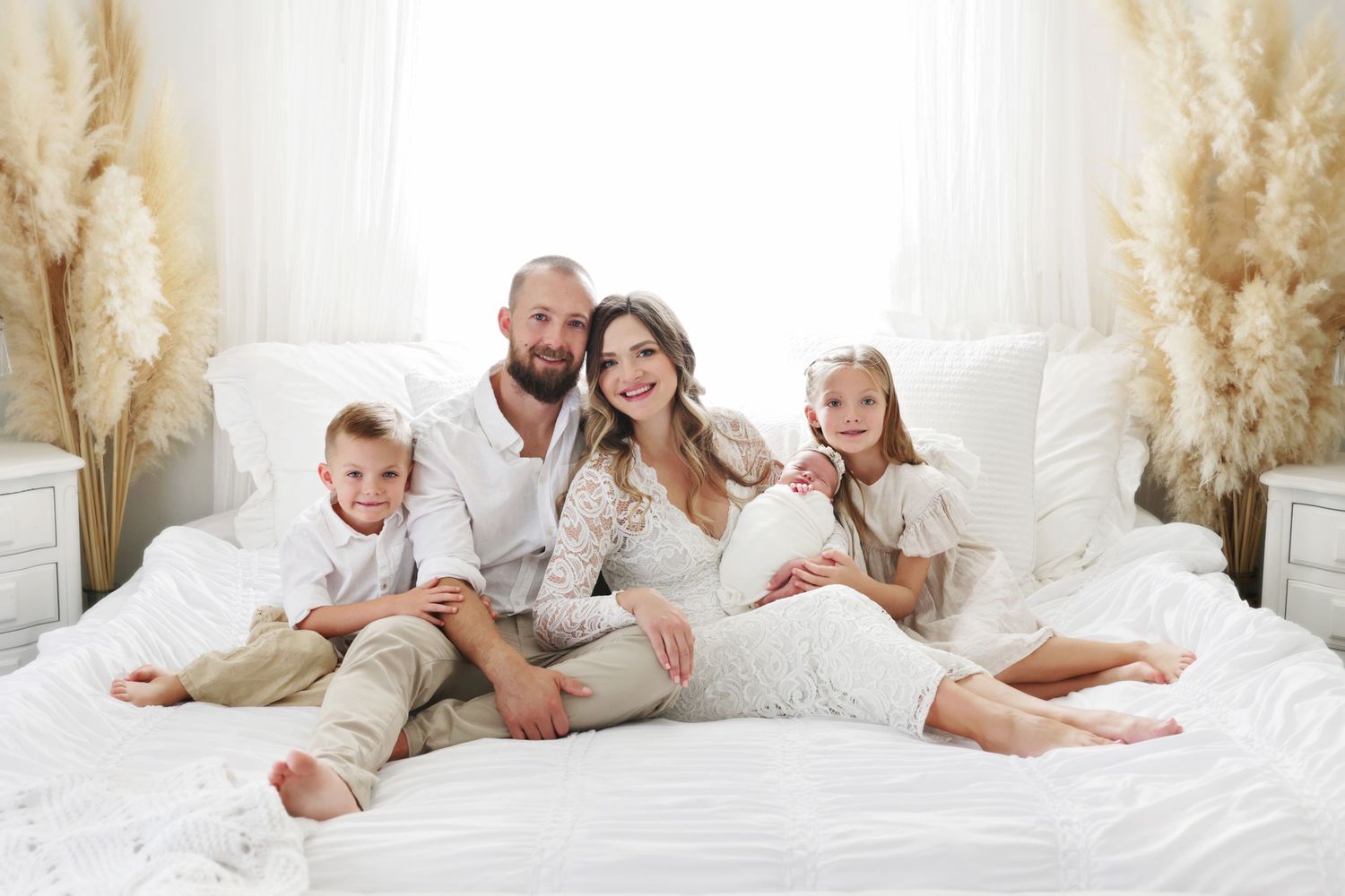 Family of four poses on white bed for maternity photo, parents and two children in matching white outfits.