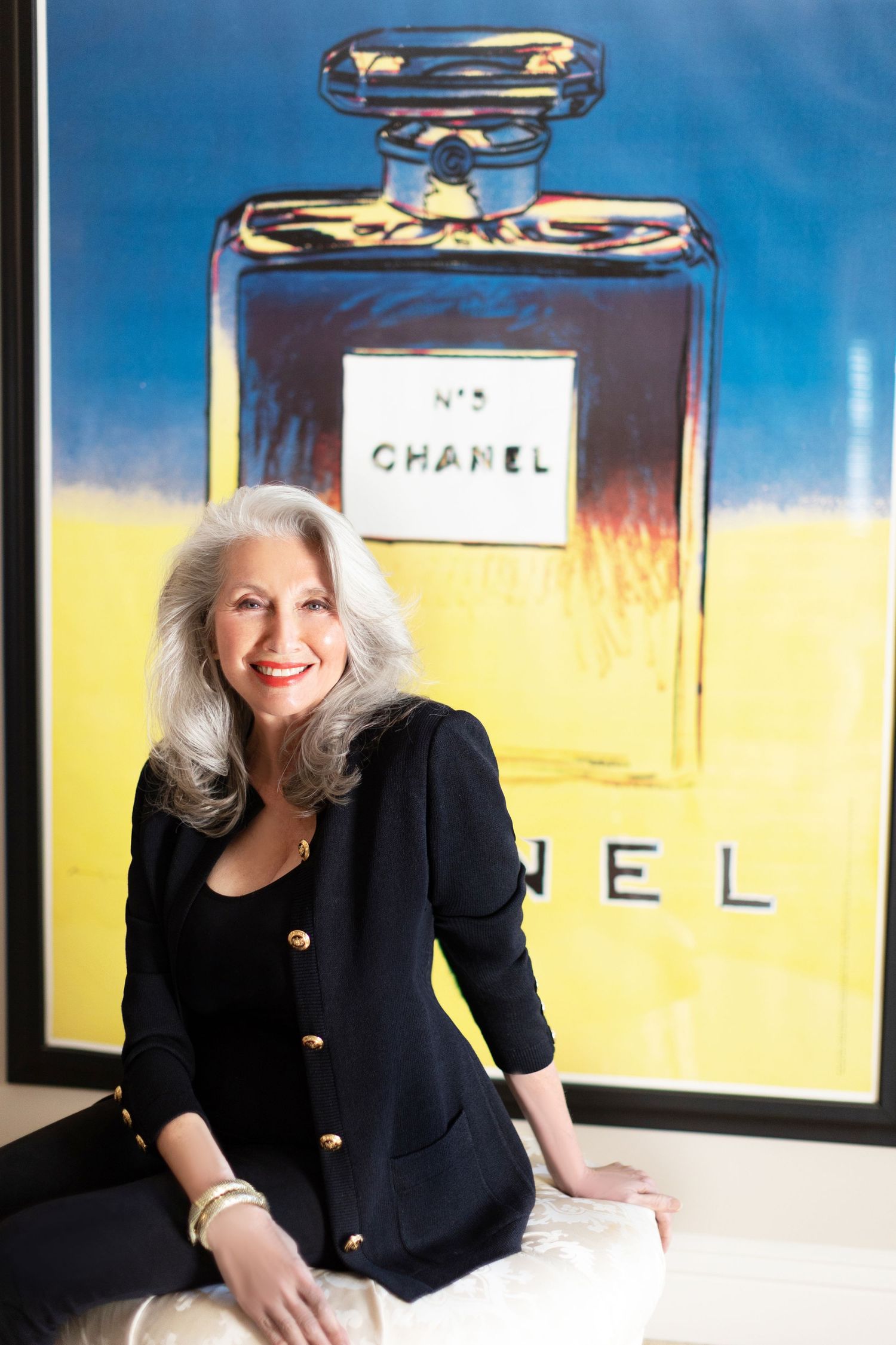 Elegant woman with silver hair in black blazer poses before Andy Warhol-style Chanel No. 5 perfume bottle artwork.