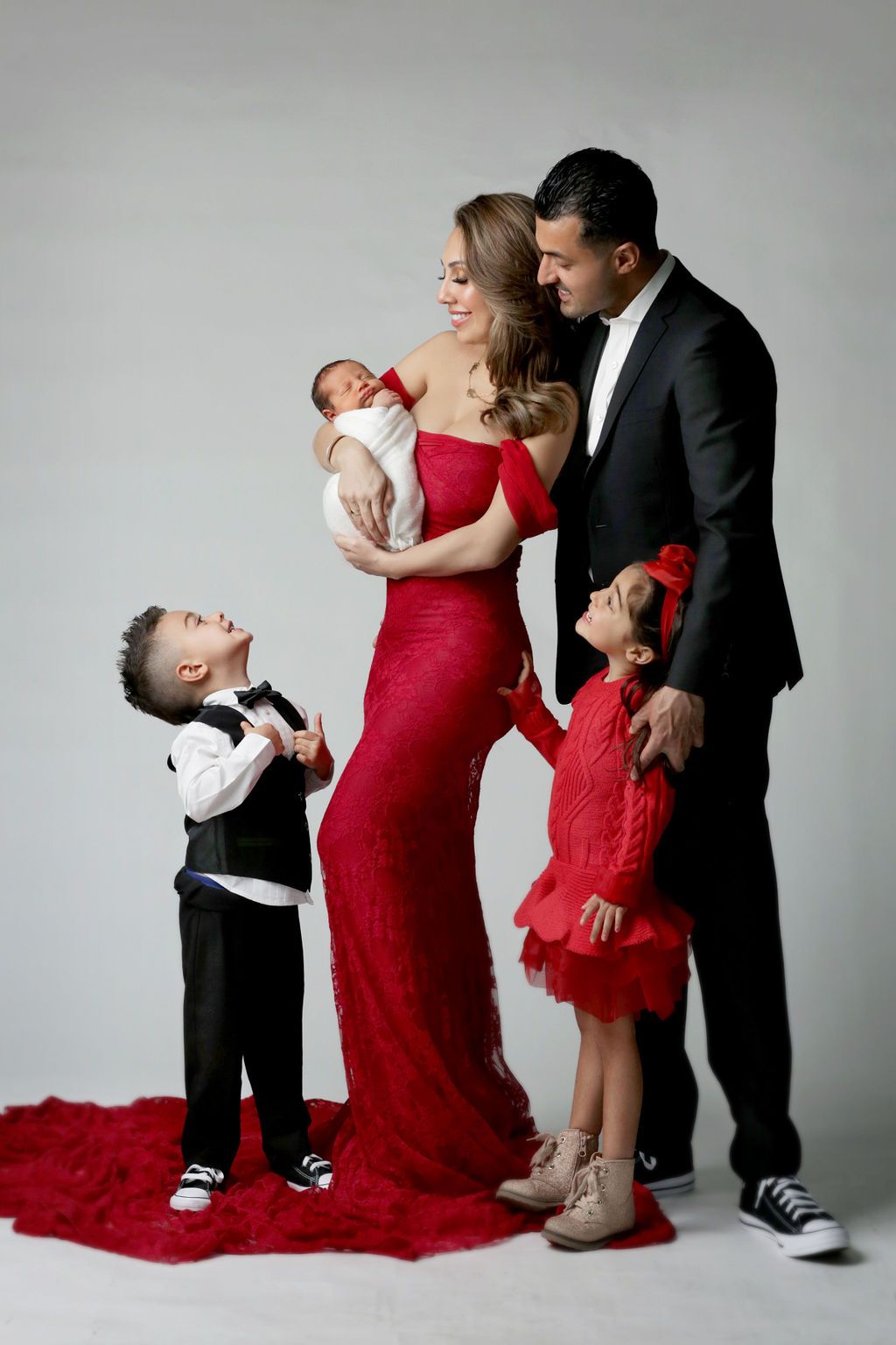 Elegant family portrait with parents in red dress and black suit holding newborn, with two young children posing.