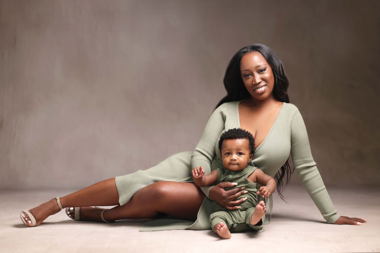 A smiling Black woman in a sage green dress poses with her baby in a matching outfit in a studio portrait.