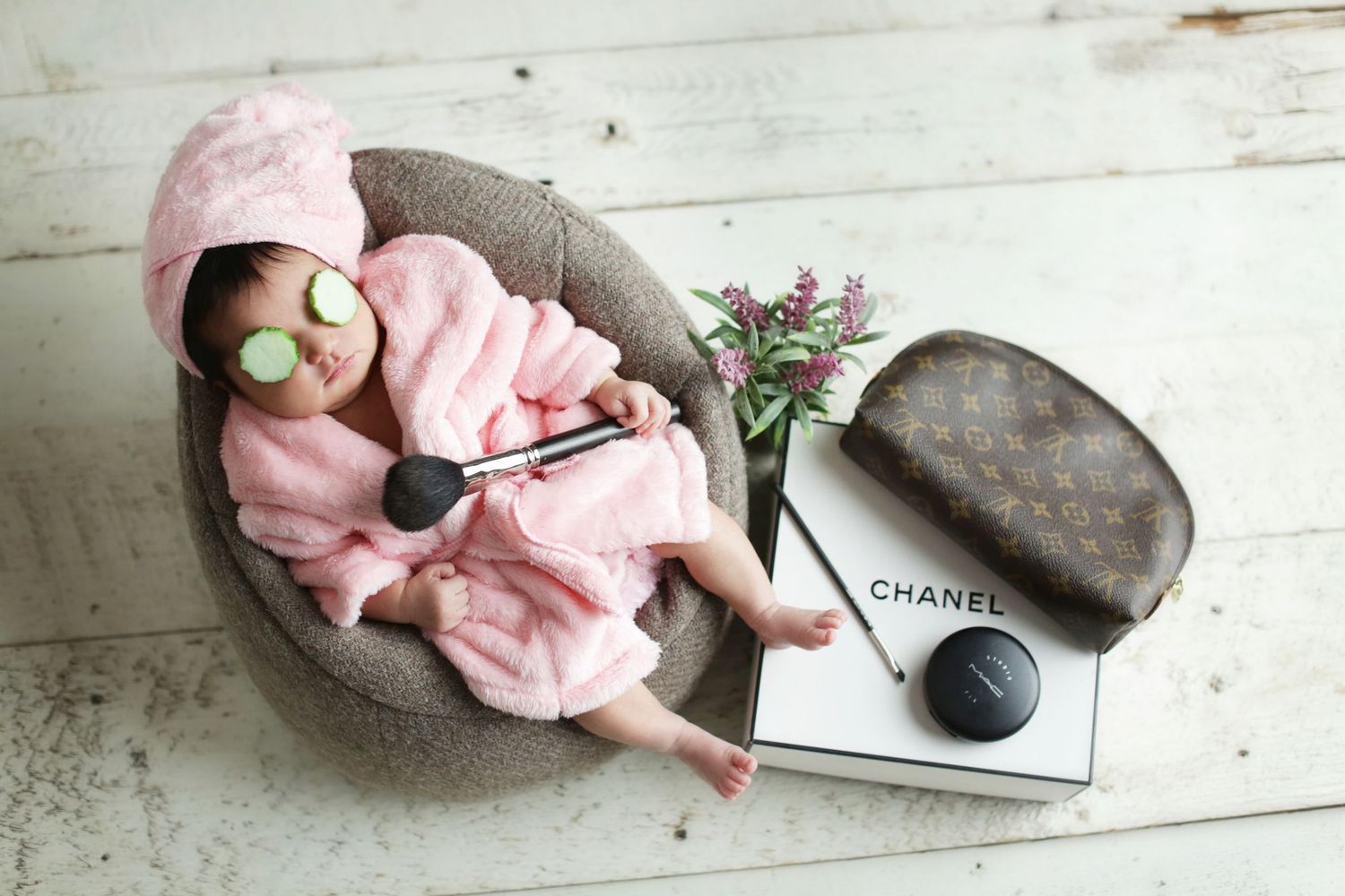 Newborn baby posed in spa theme with Chanel accessories, cucumber eyes, and pink robe for photo shoot.