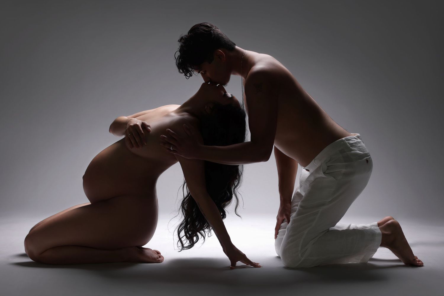 Artistic silhouette maternity photo of couple kneeling together in dramatic studio lighting.