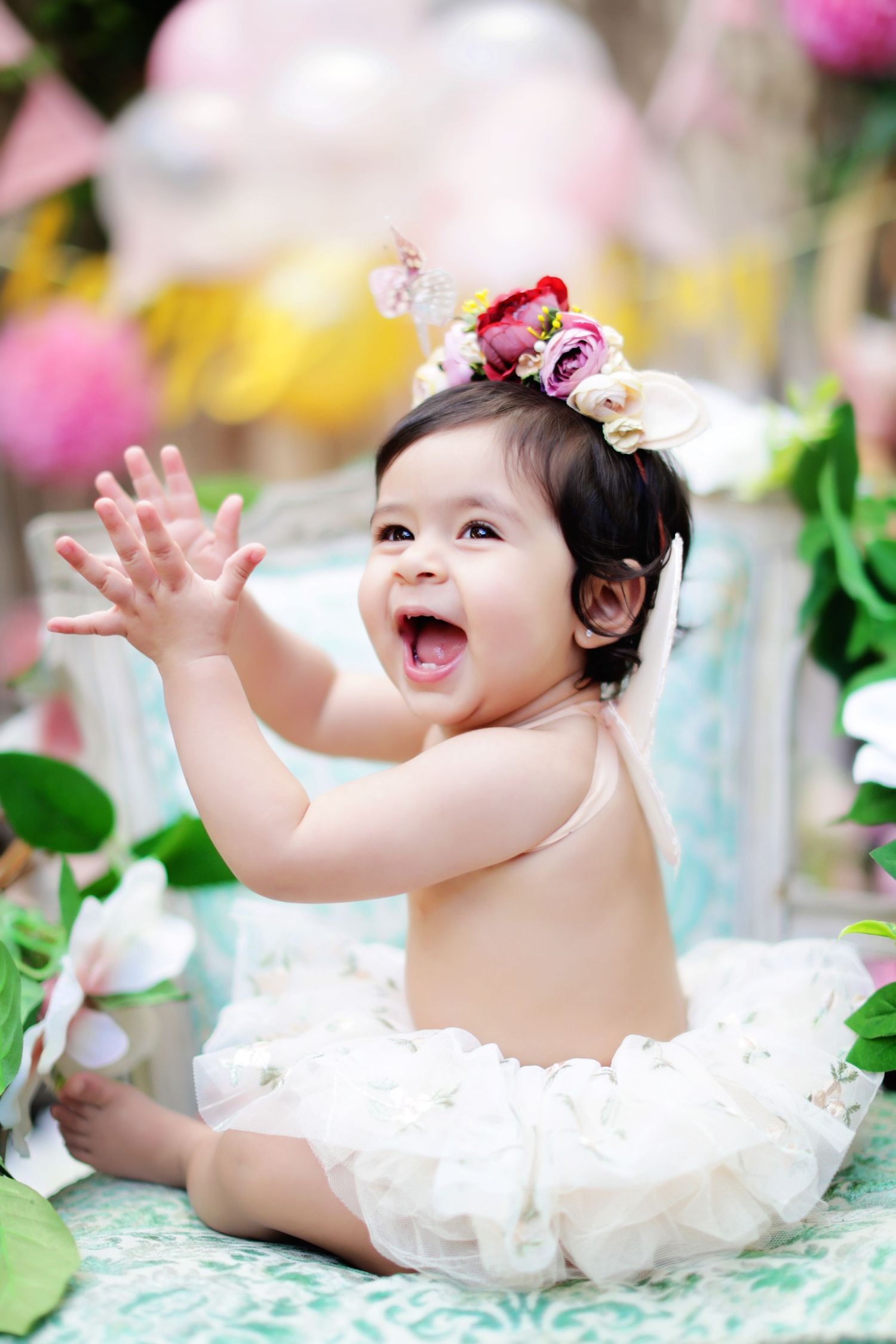 Joyful baby girl in white tutu with floral crown claps hands at outdoor birthday celebration.