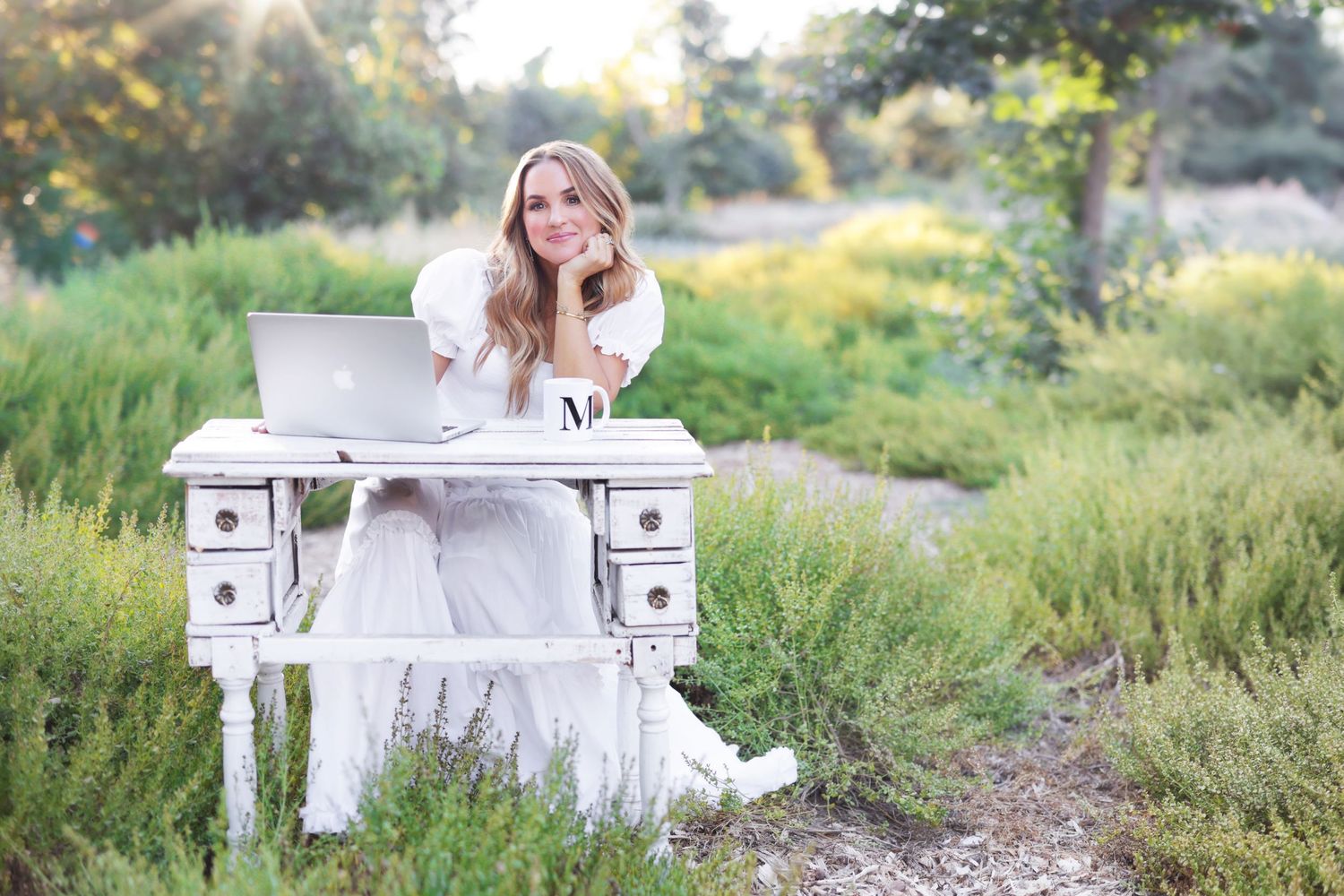 Woman in white dress sitting at vintage desk with laptop outdoors surrounded by lush green foliage.