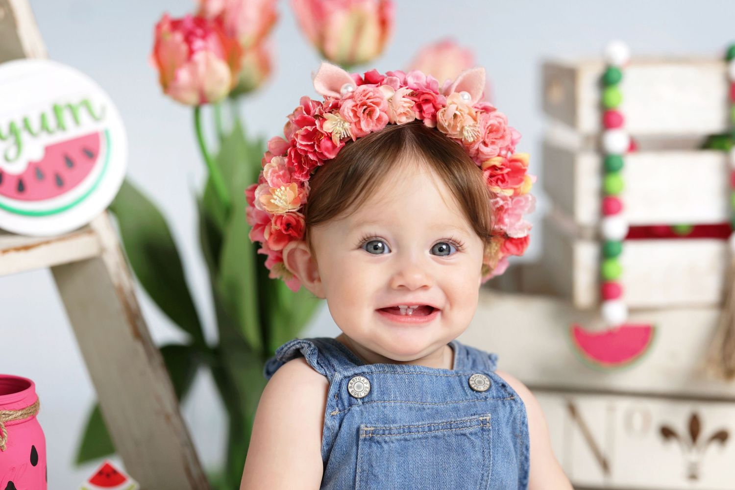 Cute baby girl in denim overalls wearing a pink floral crown smiling with watermelon-themed decorations and tulips.
