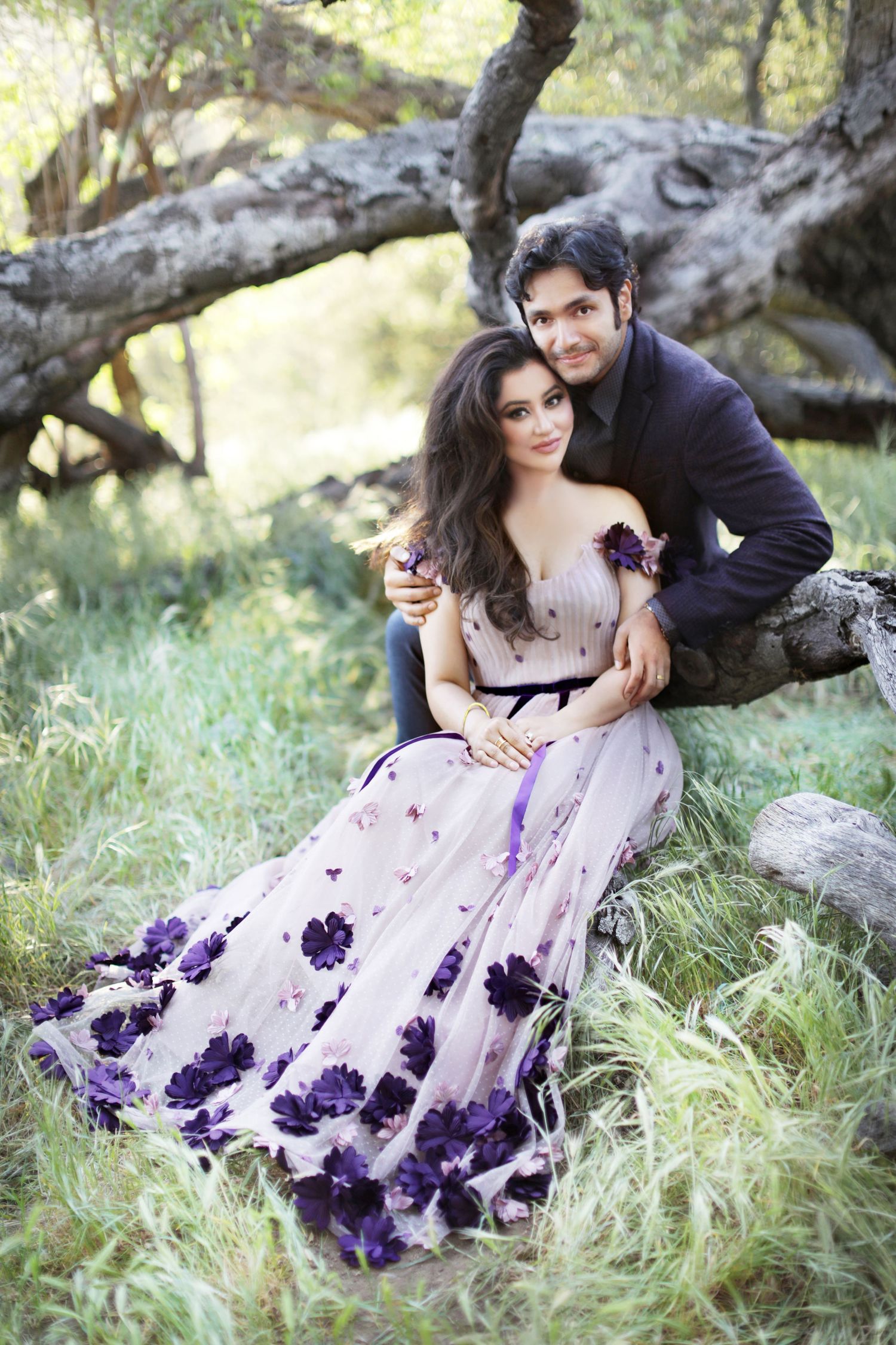 Romantic couple posing outdoors under a tree, woman in floral purple gown, man in dark suit embracing her.