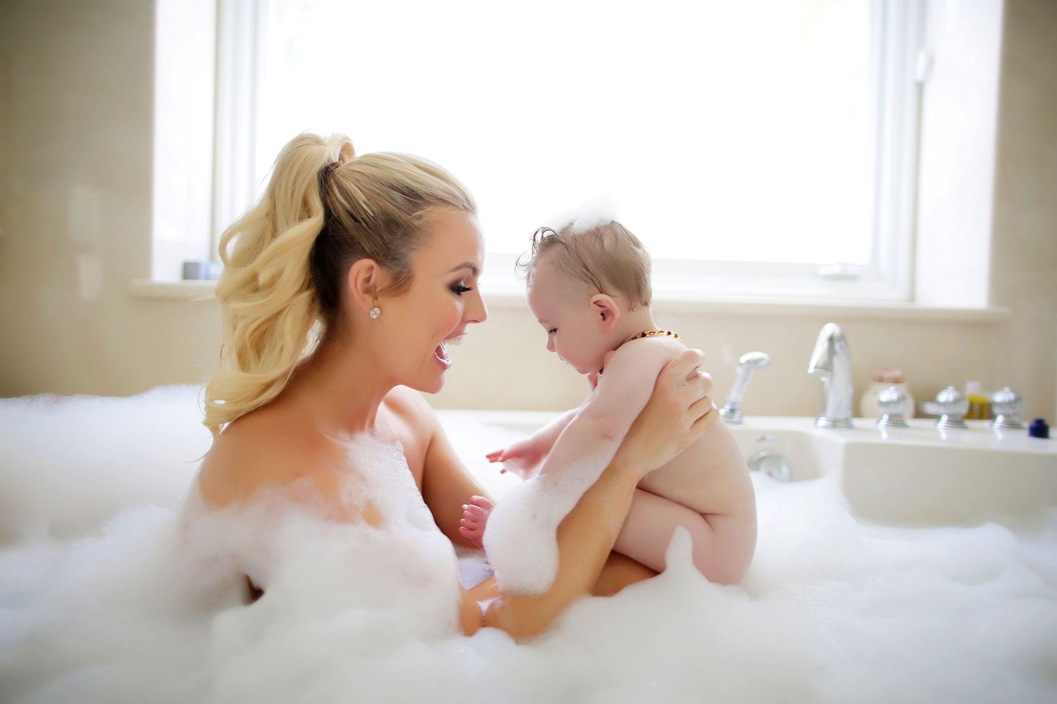 Mother and baby enjoying a bubble bath together, sharing a tender moment in a white bathtub.