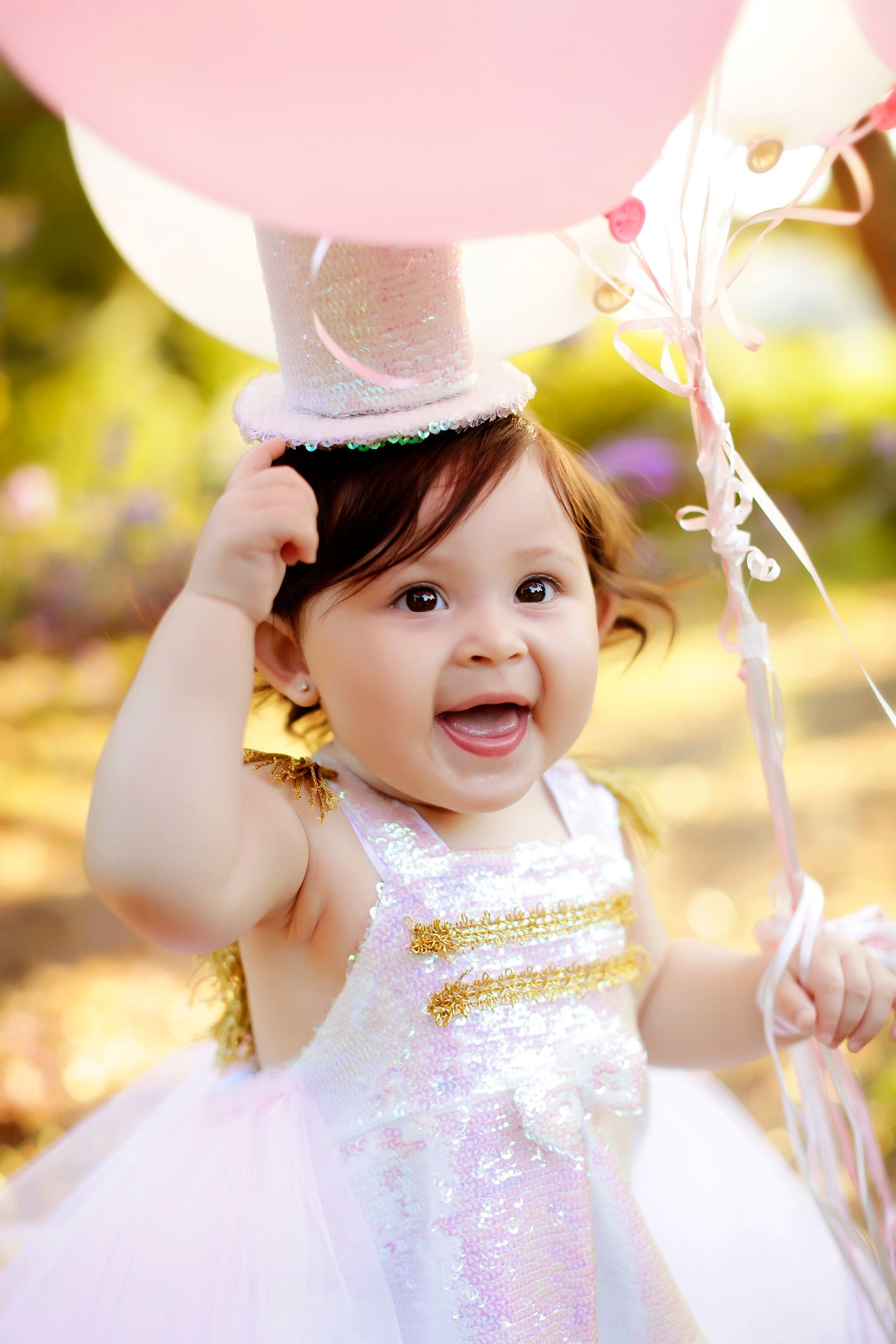 A joyful baby girl in a sparkly white dress and pink top hat holds pink balloons outdoors at a birthday celebration.