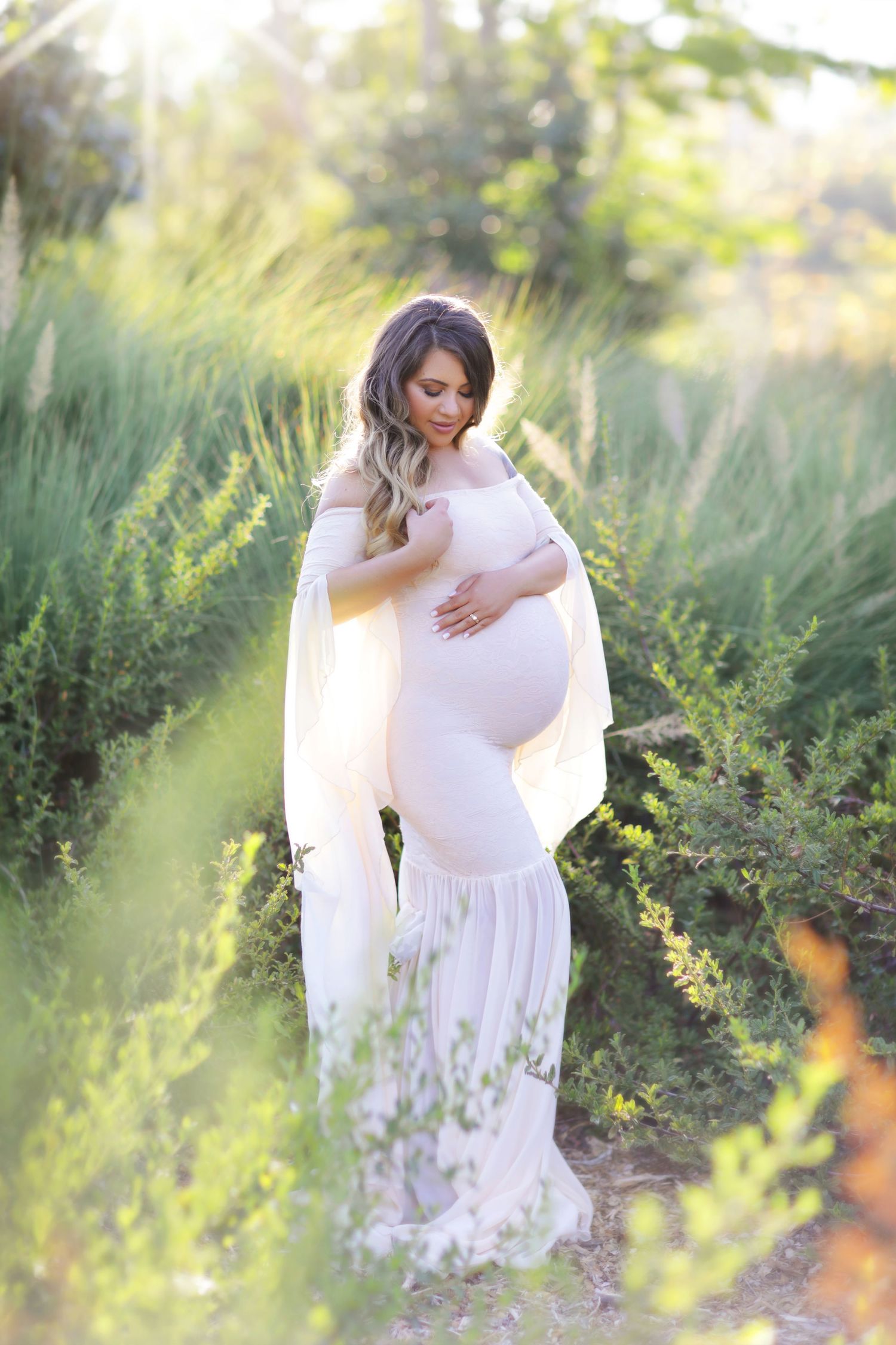 Pregnant woman in white off-shoulder gown cradling baby bump outdoors among lush green plants in golden sunlight.