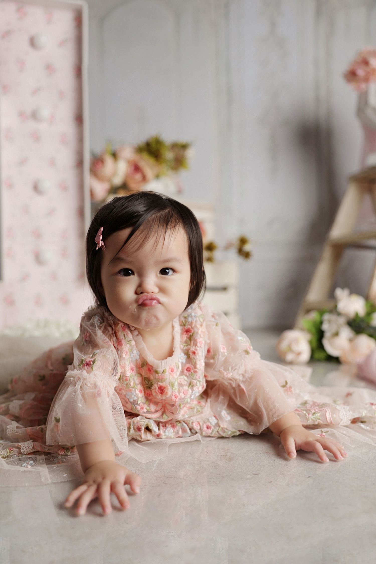Adorable baby girl in floral pink dress making a pouty face while crawling on floor in soft pastel studio setting.