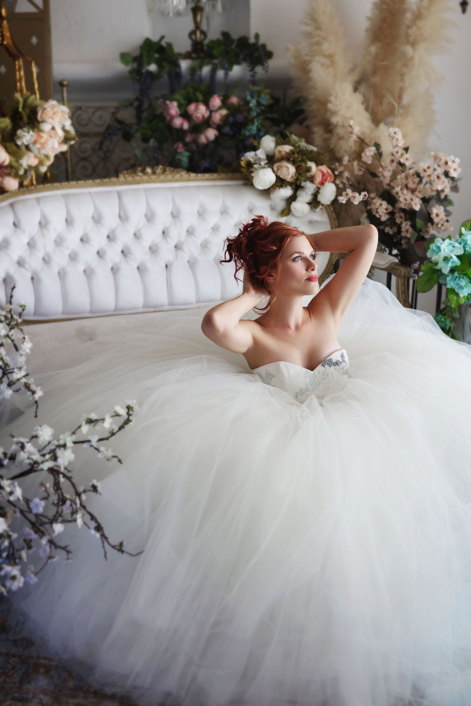 Bride in a voluminous white tulle ball gown sitting on a tufted sofa surrounded by floral arrangements.