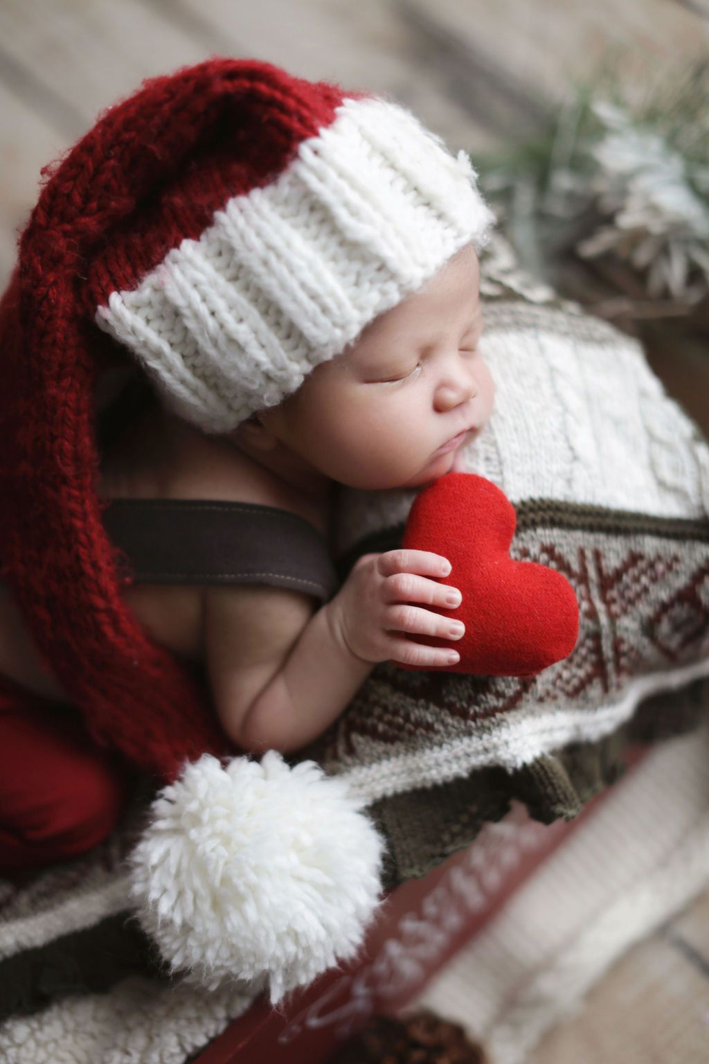 Newborn baby sleeping in Santa hat holding red heart, wrapped in cozy blanket, Christmas holiday portrait.