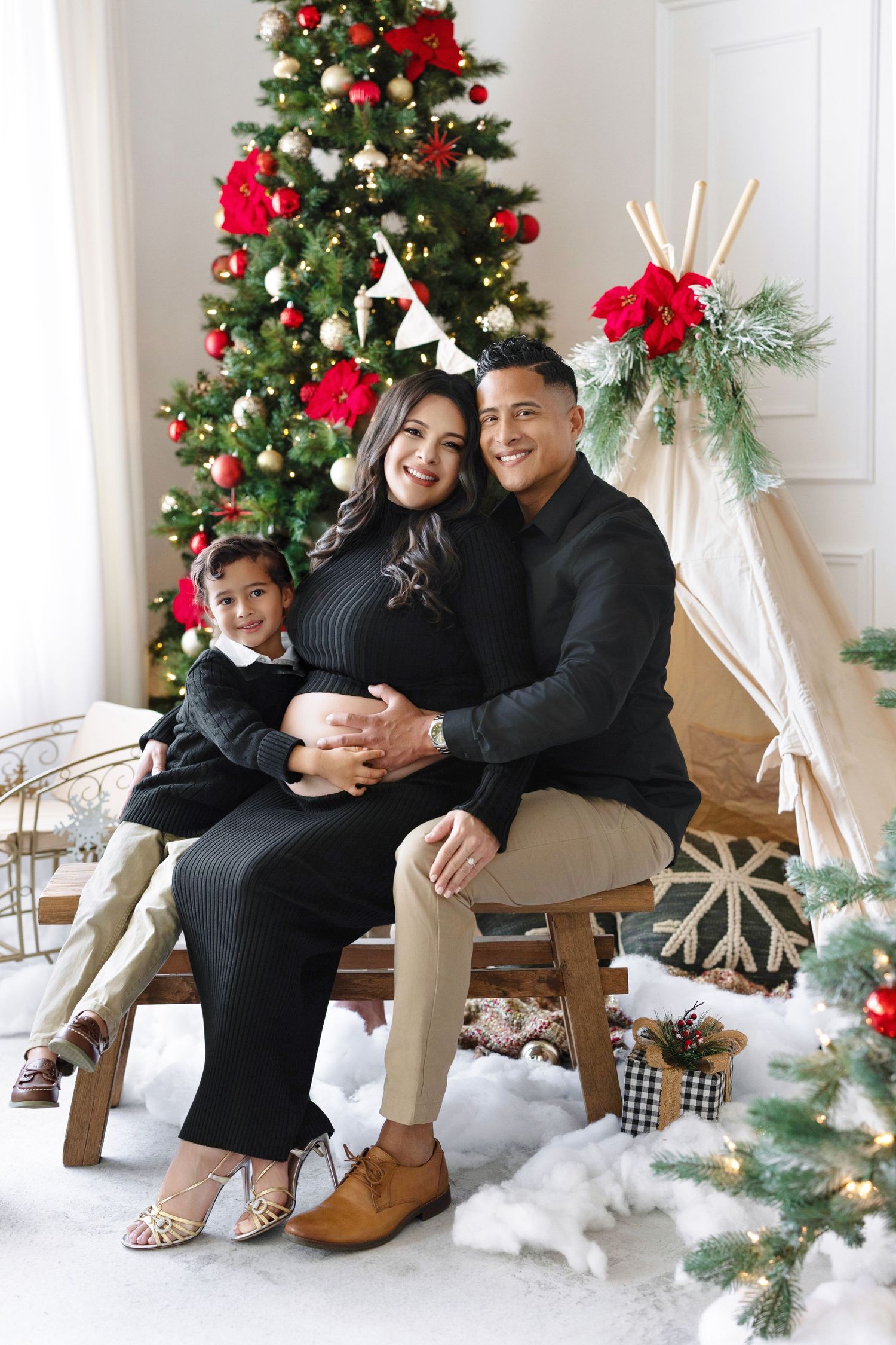Family of three in black outfits posing by decorated Christmas tree, pregnant mother seated with child and husband.