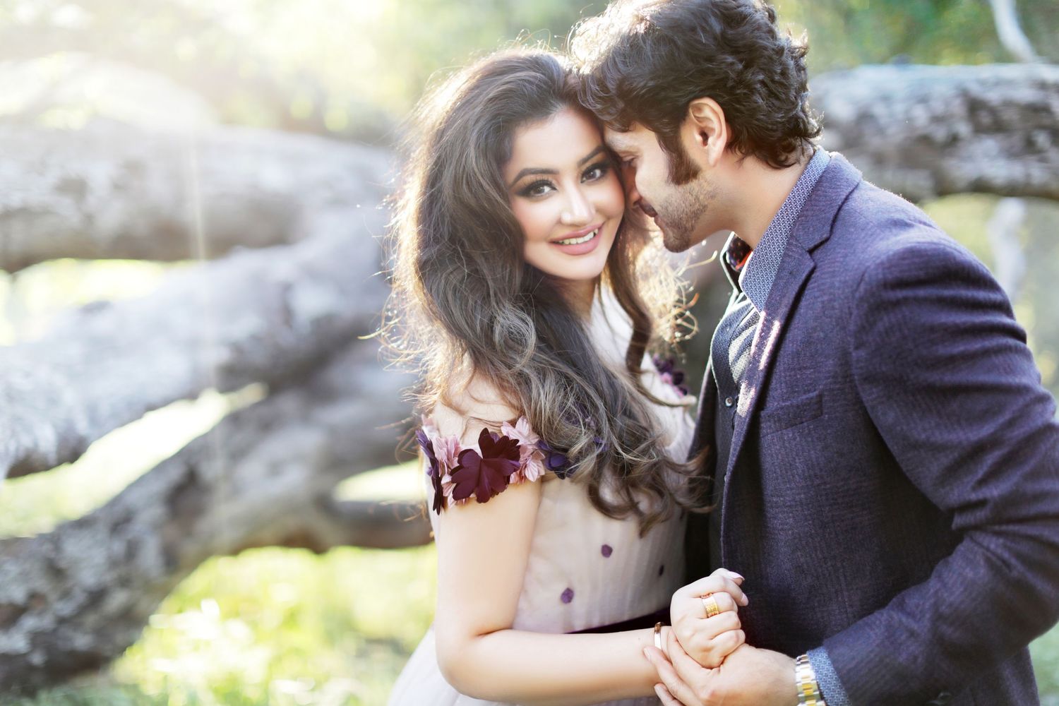 A couple in elegant attire share a romantic moment outdoors, with the woman smiling warmly at the camera.