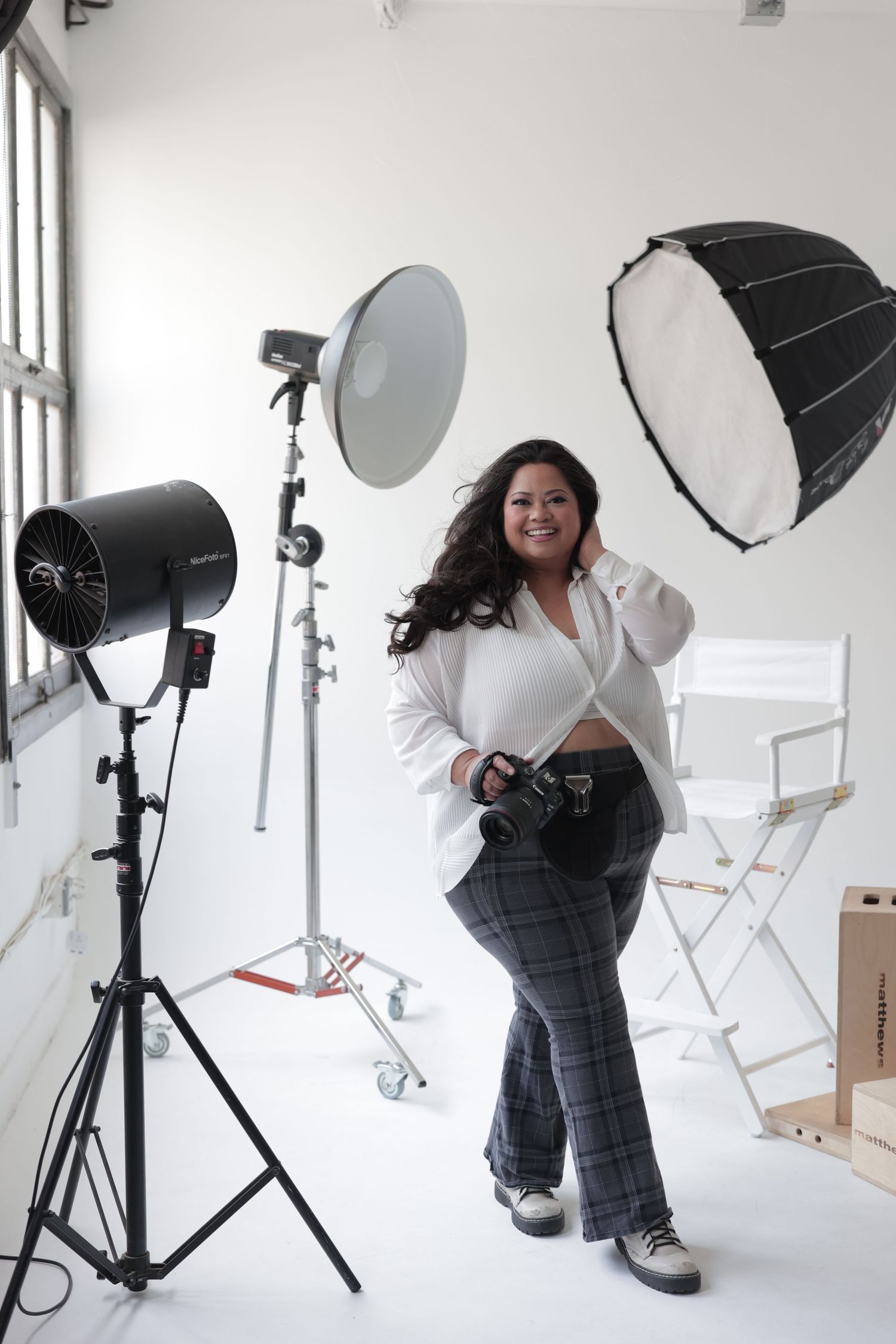 Plus-size photographer smiles holding camera in white studio surrounded by professional lighting equipment and a directors chair.