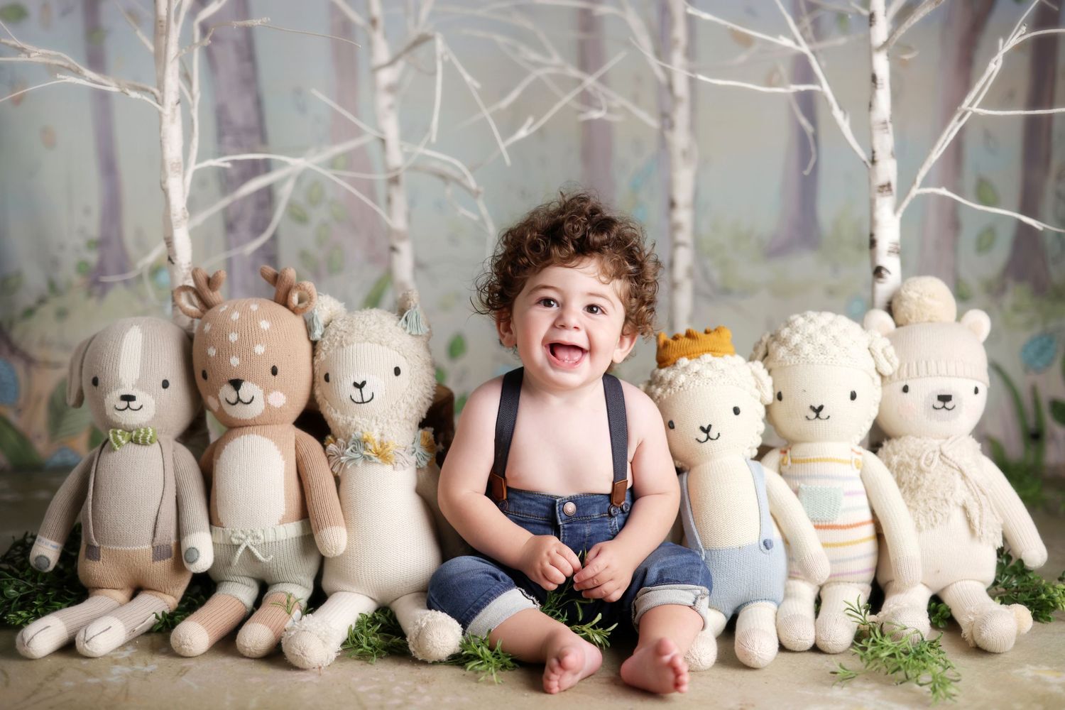 Laughing toddler boy in suspenders sitting among a row of knit stuffed animals.