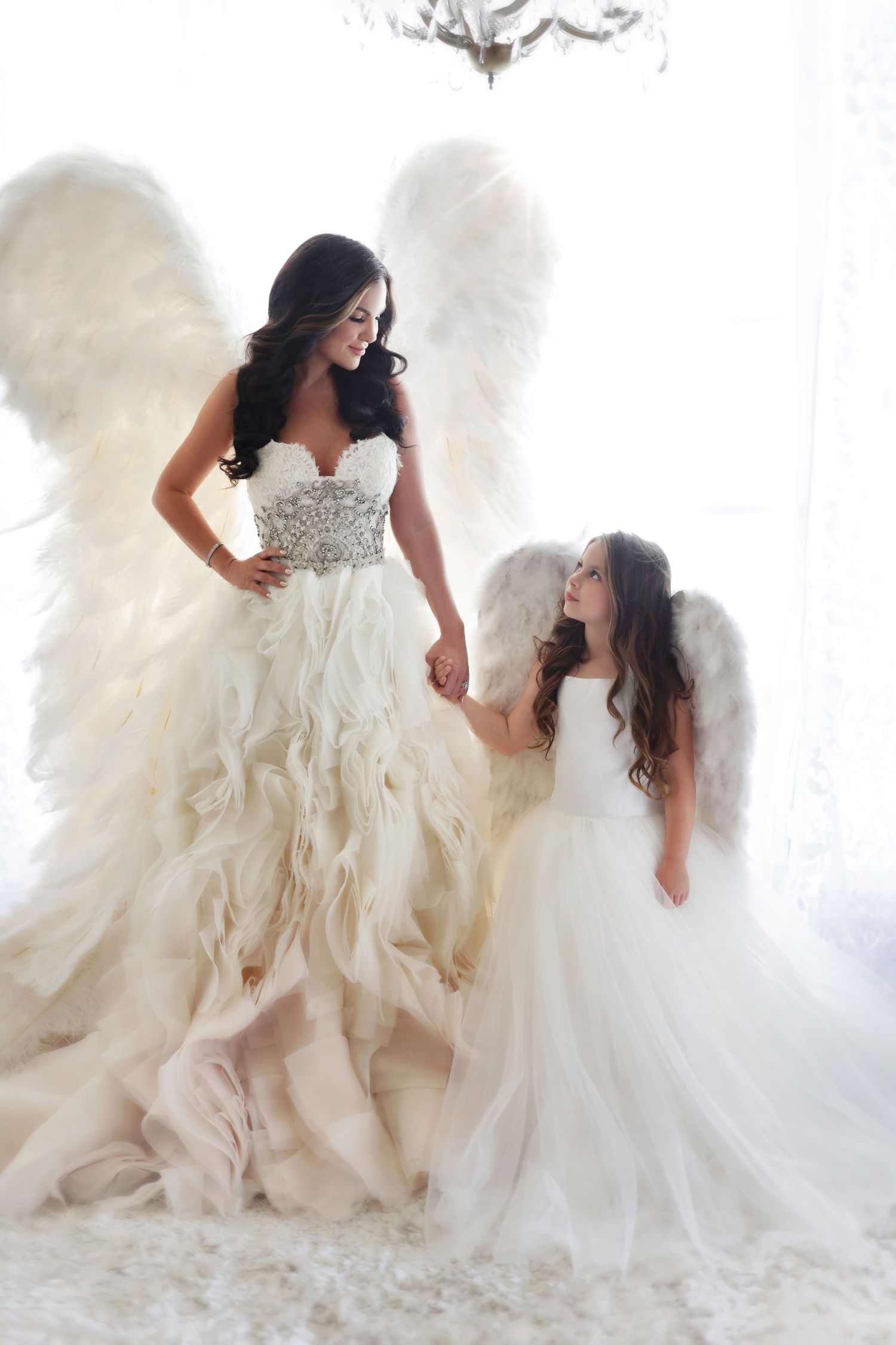 Mother and daughter in matching angel costumes with white wings and elegant gowns, sharing a tender moment.