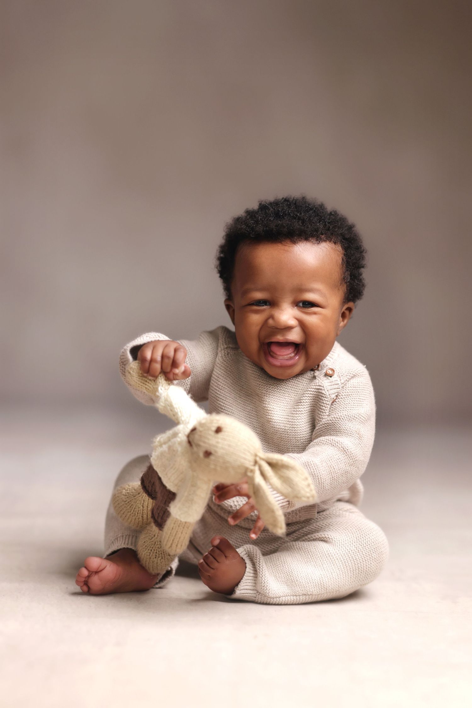 Happy baby in beige knit romper laughing while playing with stuffed bunny toy, sitting on floor.