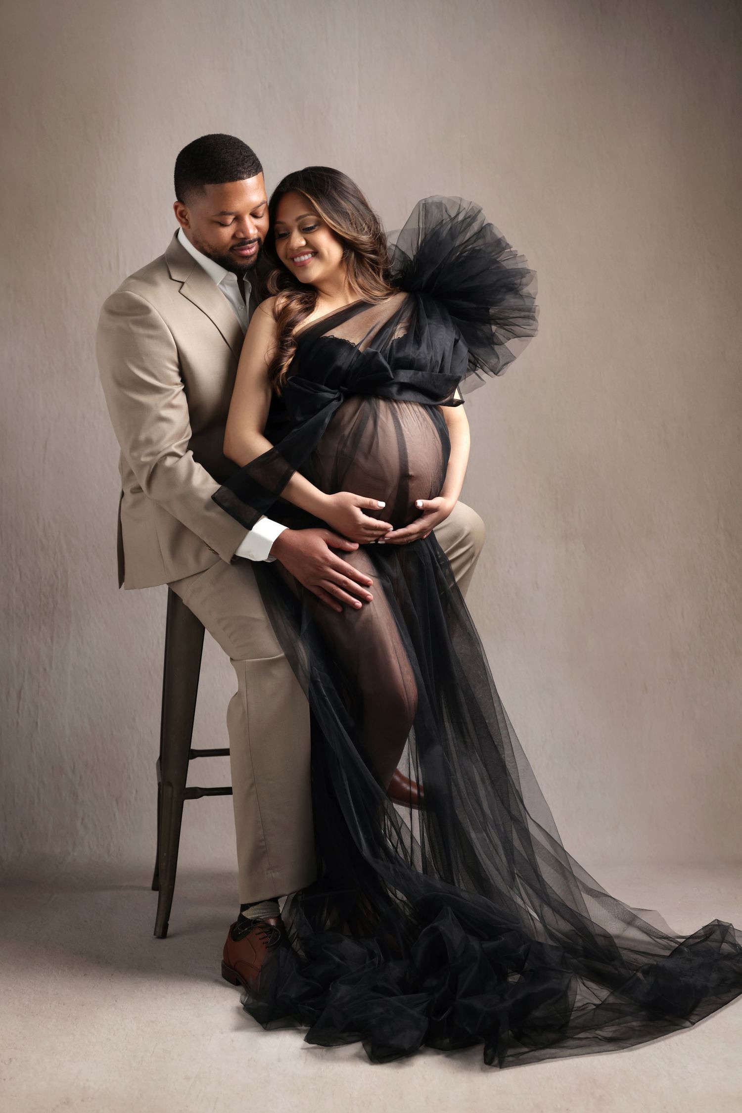 Elegant maternity photo of a pregnant woman in black tulle gown with her partner in a beige suit in a studio.