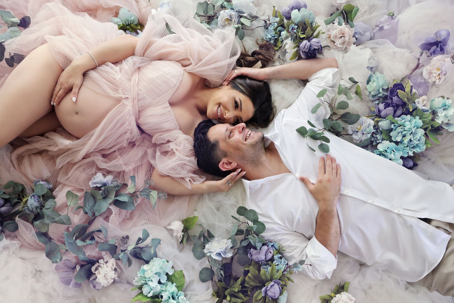 A couple lies among blue and purple flowers, the pregnant woman in a pink gown, her partner in white, gazing lovingly.