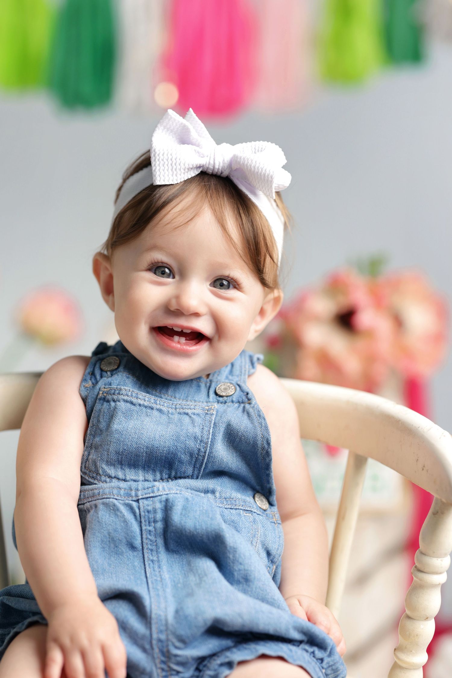 Smiling baby girl in denim overalls and white bow headband sitting in a wooden chair with colorful party decorations.