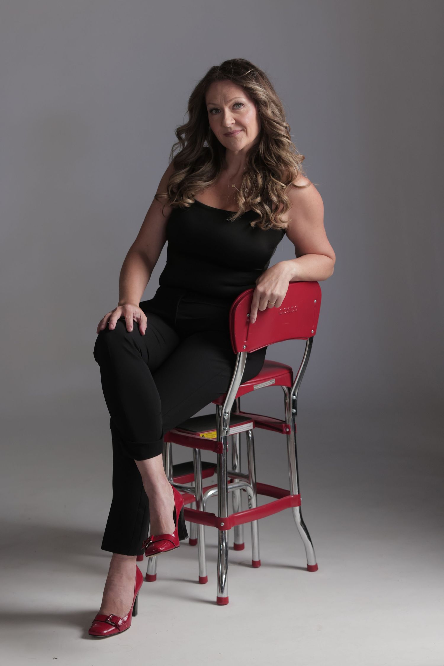A woman in a black strapless jumpsuit and red heels sits gracefully on a vintage red chrome step stool chair.