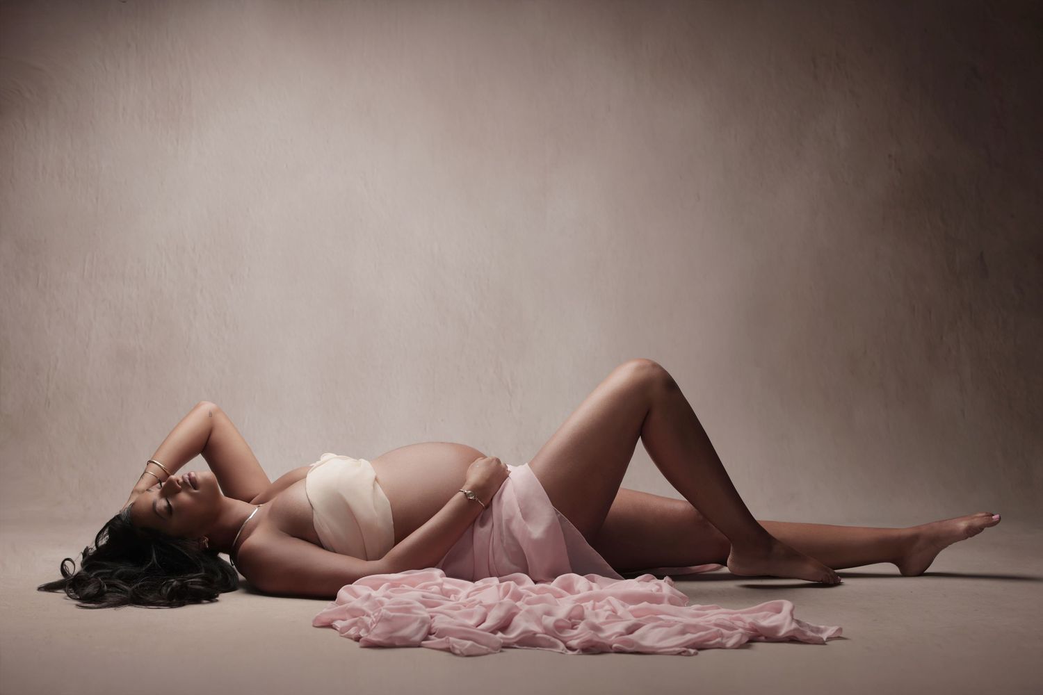Pregnant woman in pink lingerie lying gracefully on studio floor with soft pink fabric draped nearby.