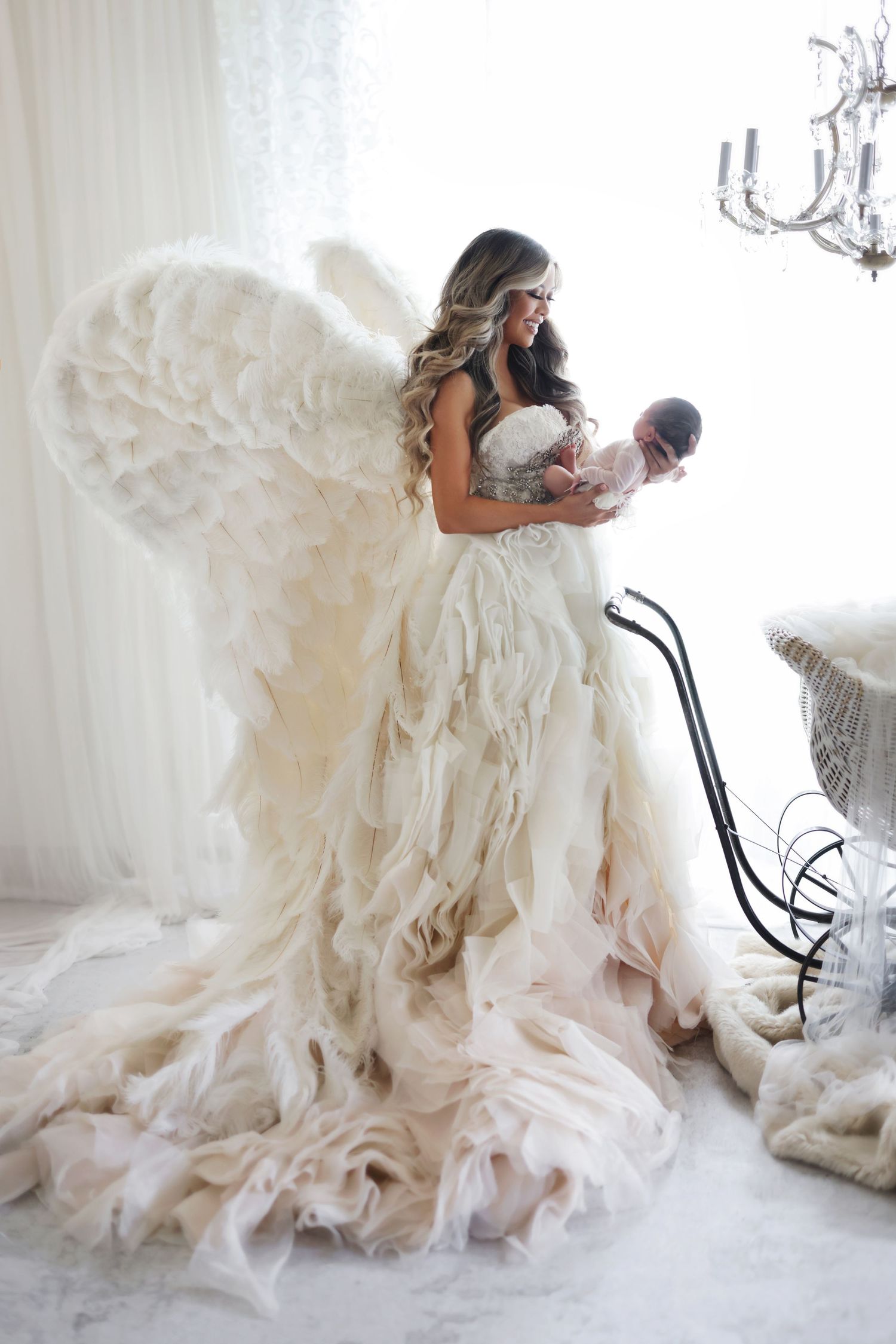Woman dressed in flowing ruffled gown with large feathered angel wings cradling a newborn indoors.