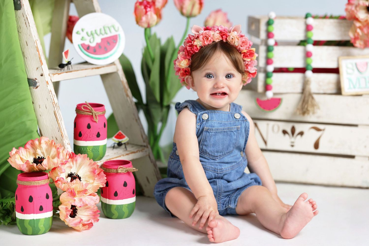 Adorable baby girl in denim overalls wearing a pink floral crown sitting near watermelon-themed decorations and flowers.
