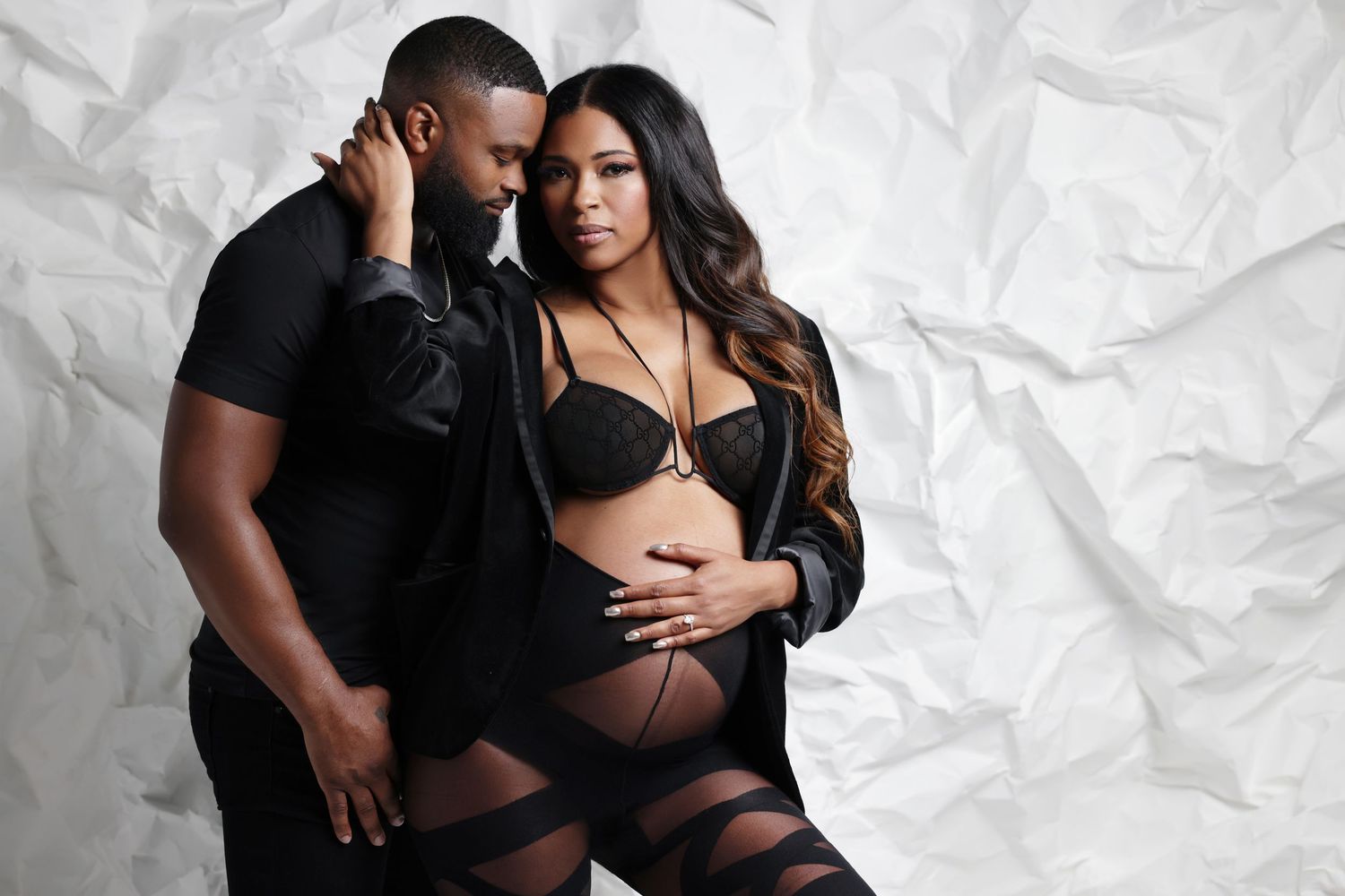 Couple maternity photo shoot in black outfits against white backdrop, woman in lingerie cradling pregnant belly.