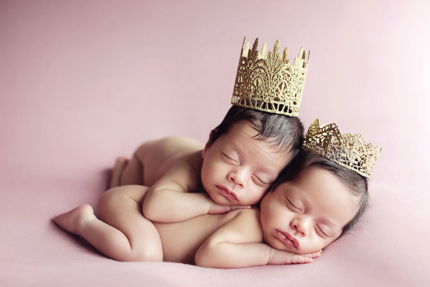 Two sleeping newborn twins wearing gold crowns on a pink background.