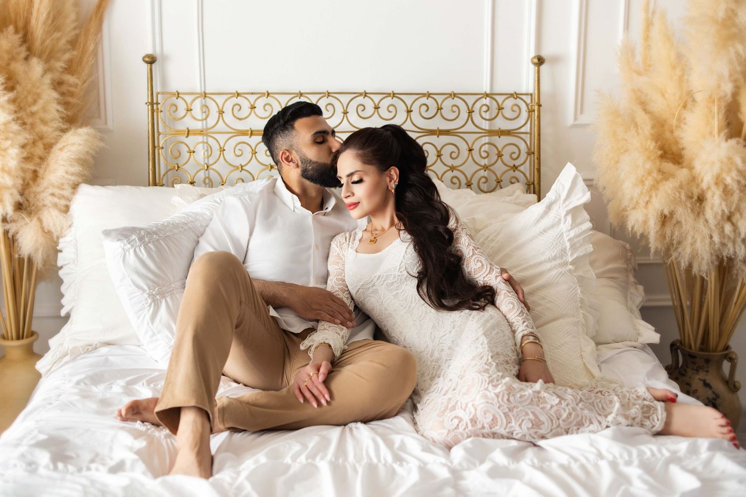 Romantic couple sitting on a white bed surrounded by pampas grass in an elegant gold and white bedroom.