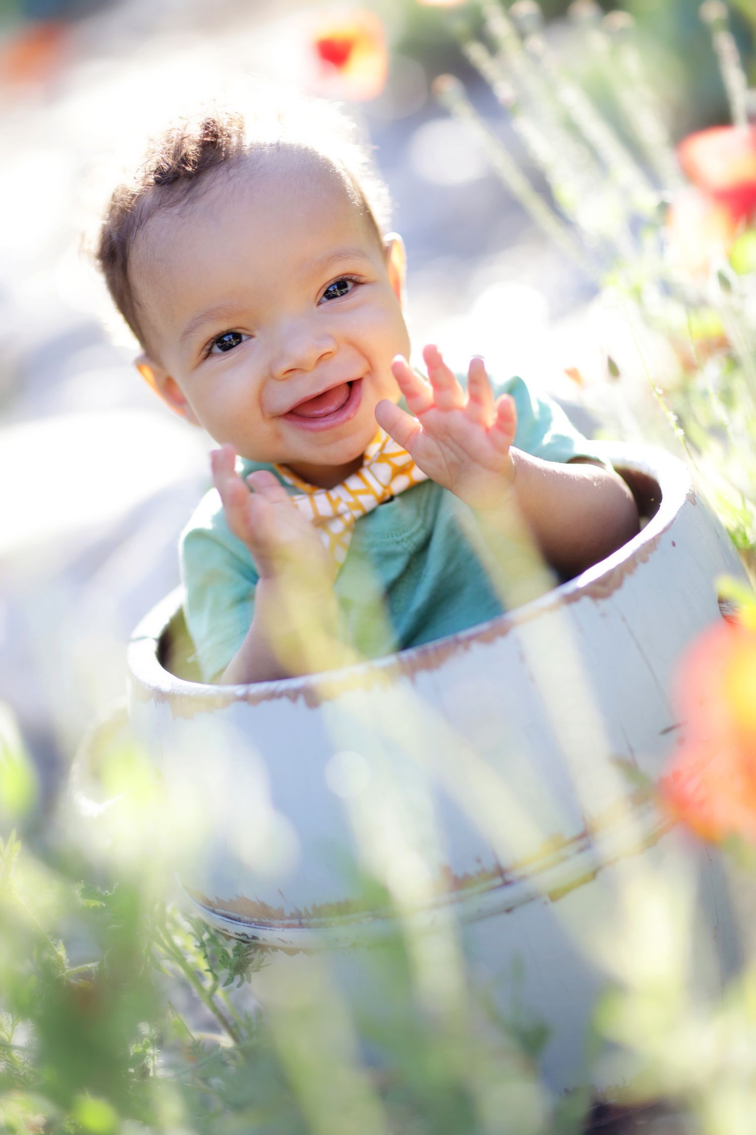 Smiling baby sitting in a white bucket planter surrounded by colorful flowers in a sunny garden.