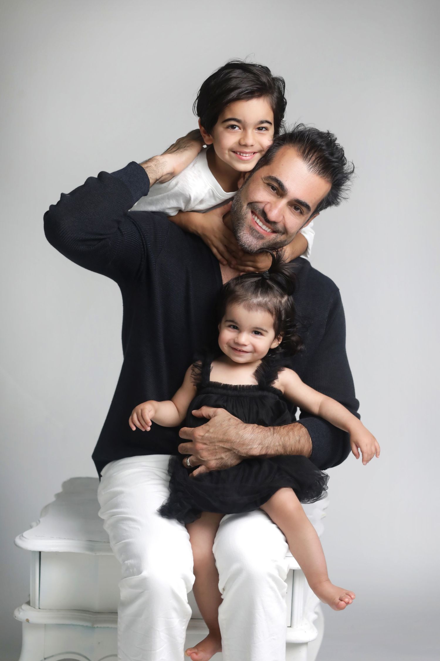 A smiling man in black sweater sits with two young daughters, one on his lap and one hugging from behind.