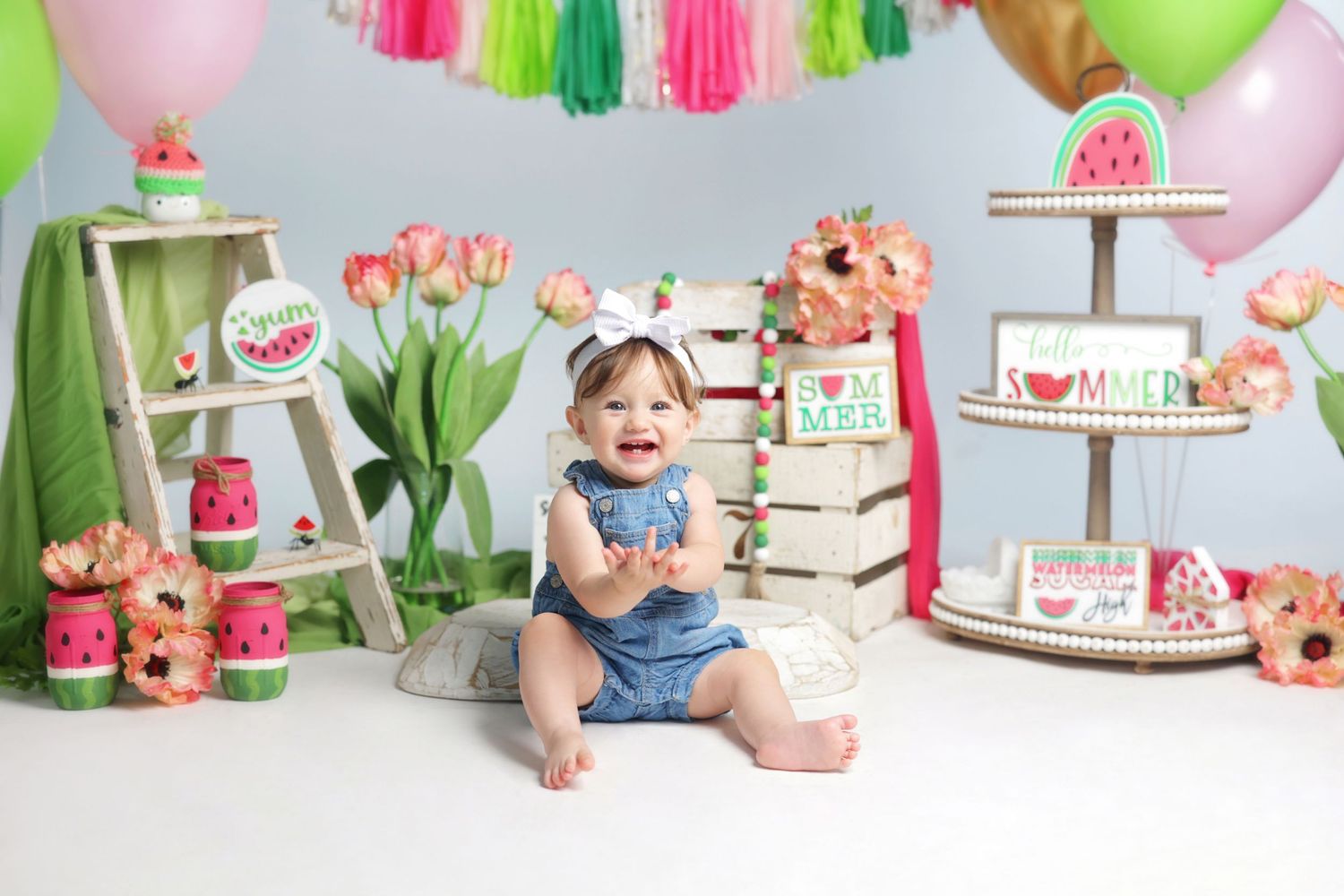 Baby girl smiles at watermelon-themed first birthday cake smash with colorful decorations, balloons, and tiered cake display.