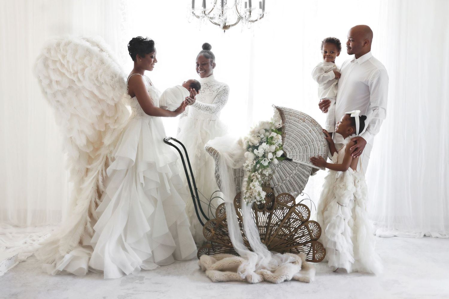 A Black family in white angelic outfits poses with feather wings and a vintage wicker baby carriage in a studio.