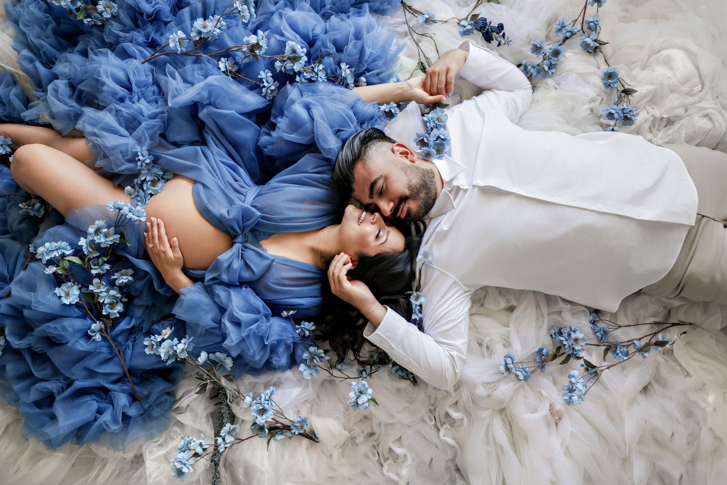 Couple lying among blue tulle fabric and forget-me-not flowers in romantic maternity photo shoot.