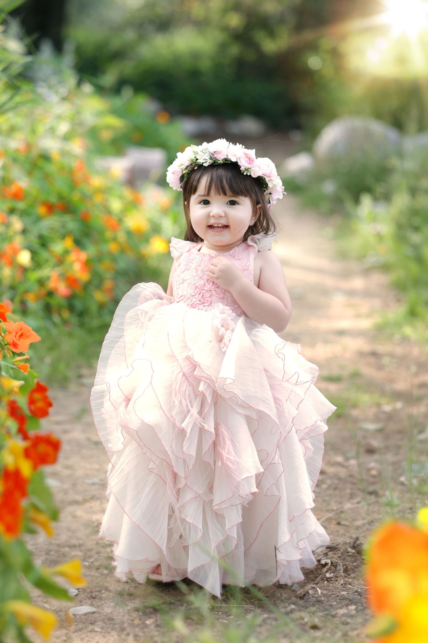Smiling toddler girl wearing pink layered dress and flower crown strolling through vibrant garden.